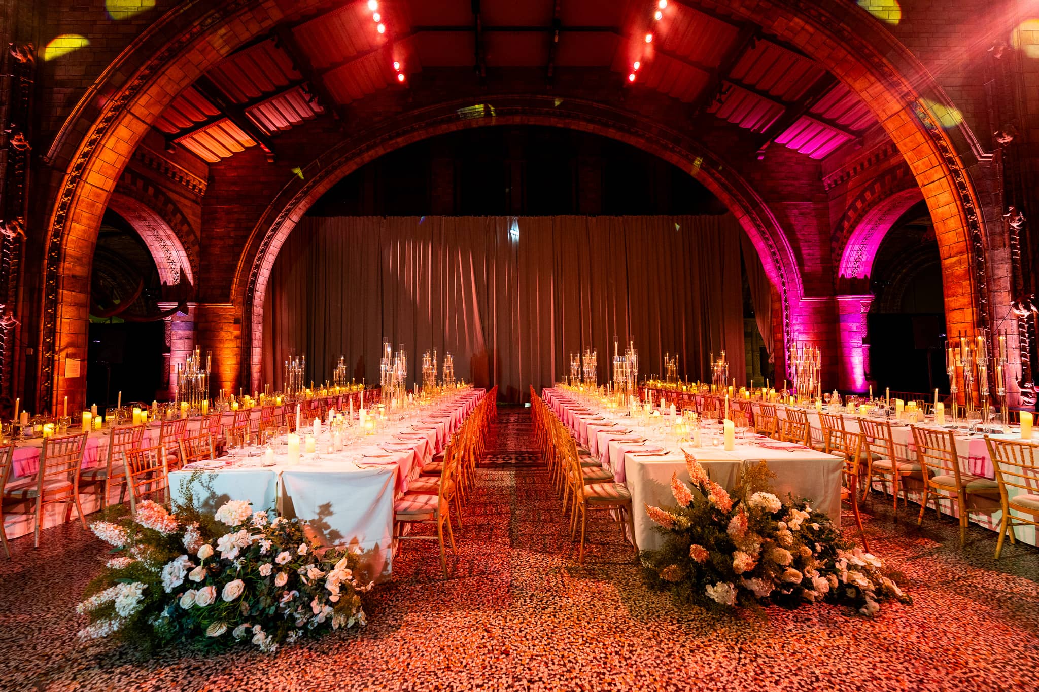 Long tables, white linen and candles under red and magenta uplighting on the Hintze Hall arches, at a Natural History Museum wedding by Kudos AV.
