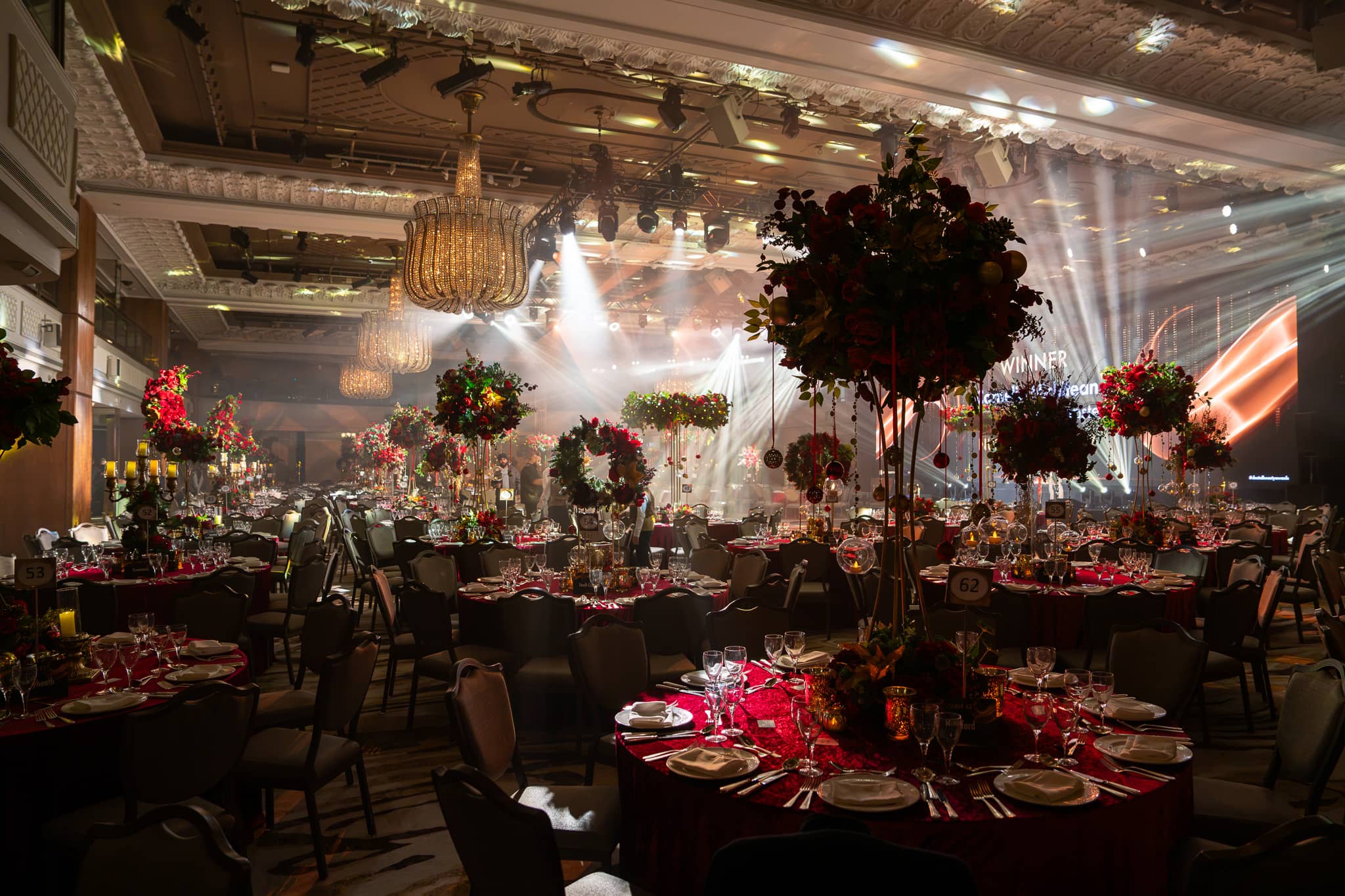 Round tables in red linen under hanging floral installations and warm light, the Dental Beauty Partners Awards 2025 dinner in the Great Room at Grosvenor House London, lit by Kudos AV.