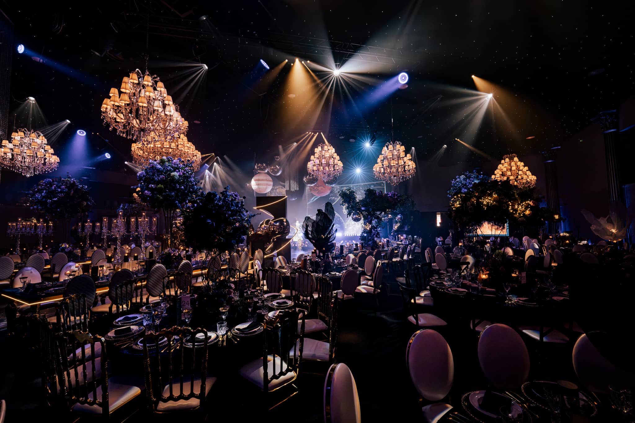 Round tables under the chandeliers with the stage and its screens lit behind, by Kudos AV