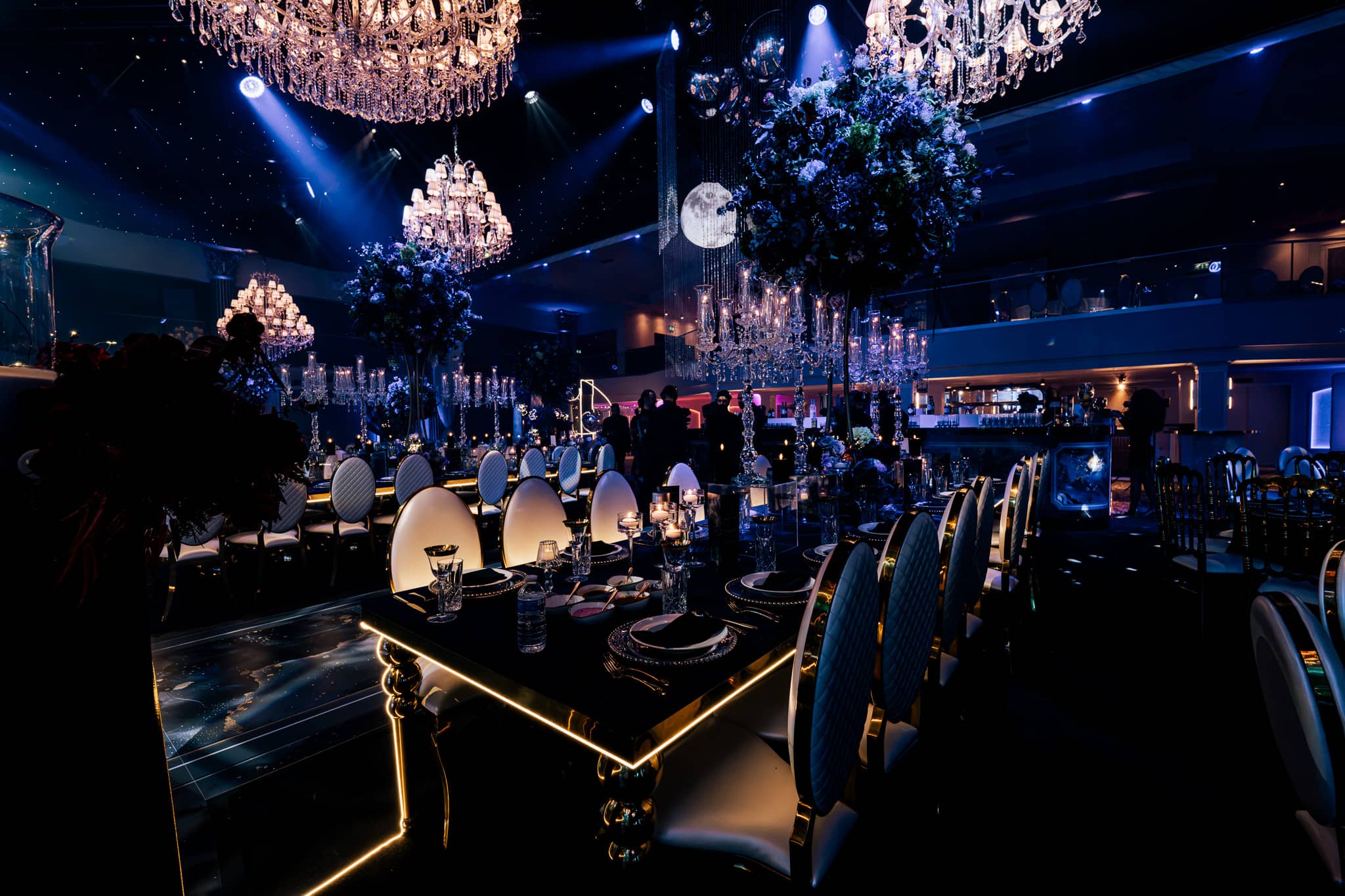 Black tables with gold edging under chandeliers in blue ambient light, by Kudos AV