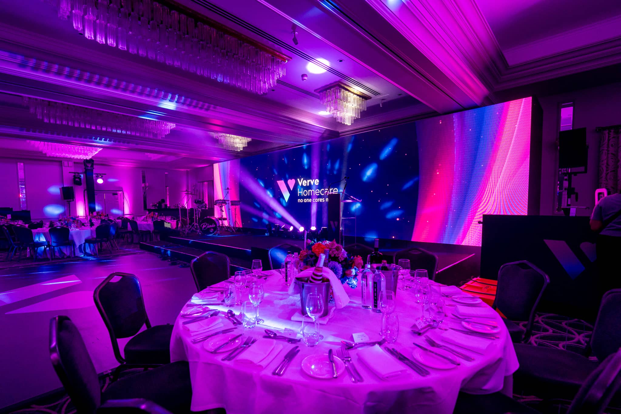 A dinner table set under magenta uplighting, with the Verve Homecare LED wall and stage beyond, in the St Alban Suite at Sopwell House.