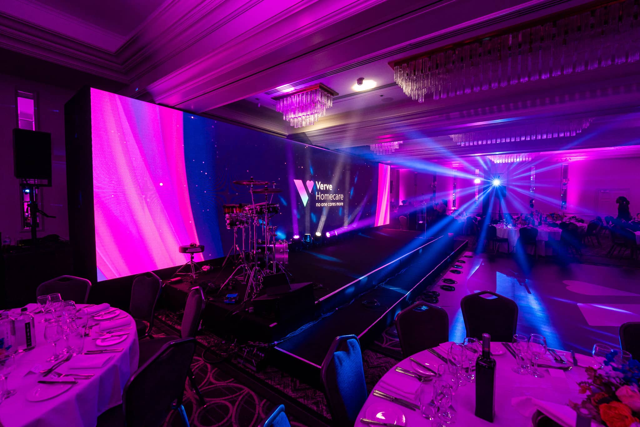 Round tables dressed for dinner under magenta light, with a beam across the room towards the LED wall at Verve Awards Night, Sopwell House.