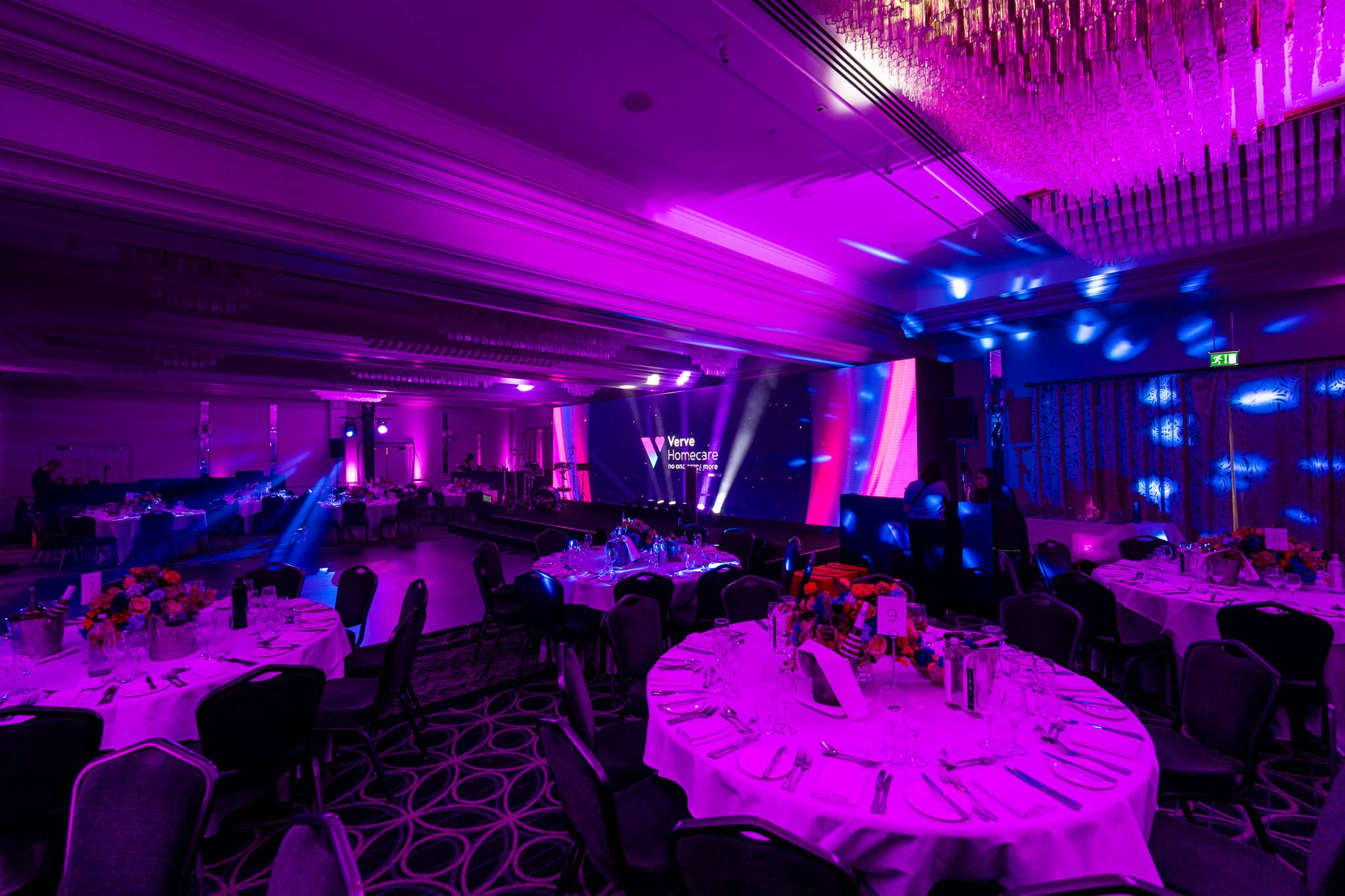 Round tables with floral centrepieces fill the St Alban Suite under magenta and blue uplighting, with beams towards the stage at Verve Awards Night.
