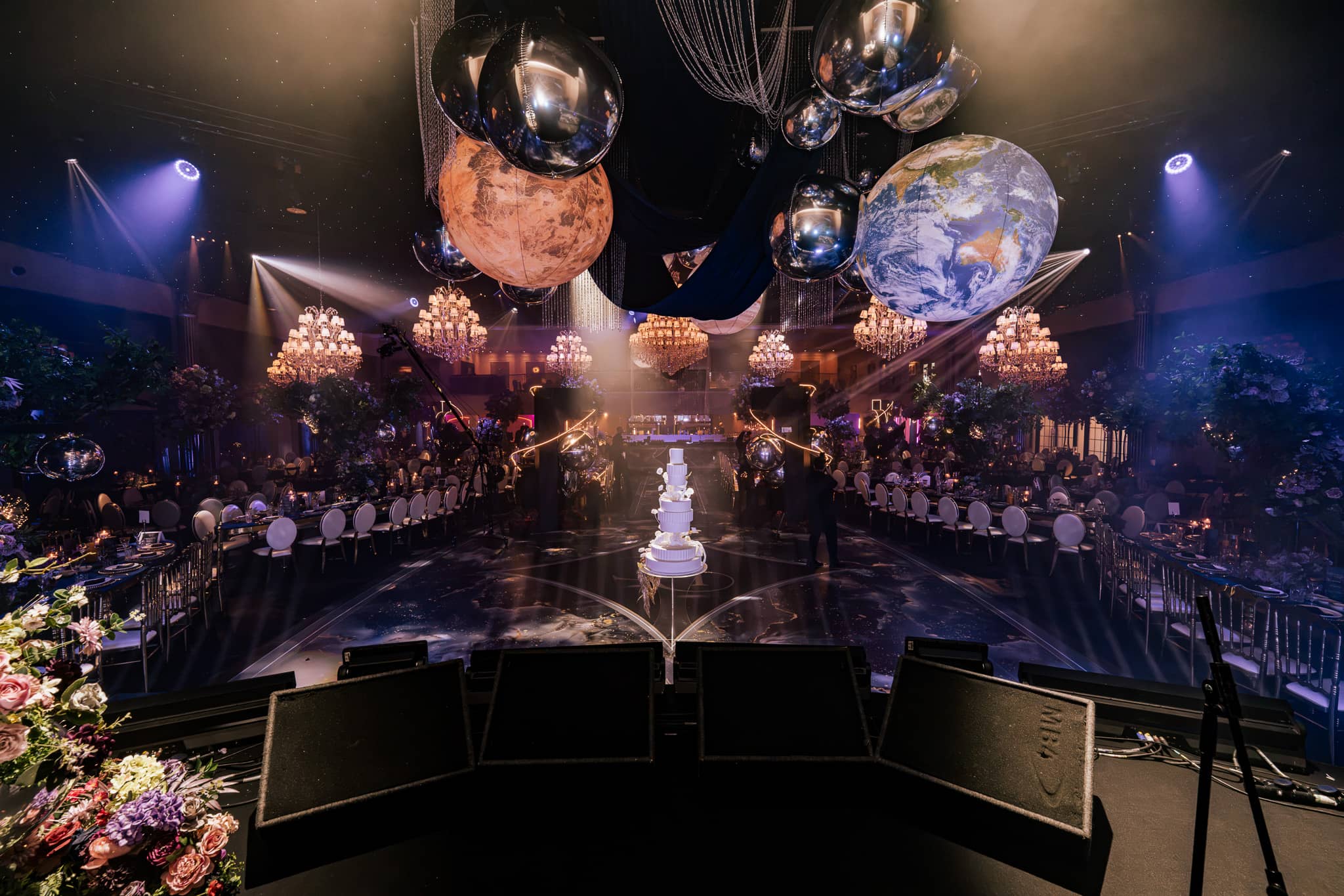 Planets suspended over the tables and the wedding cake under the chandeliers, styled by Krystal Wedding Decor and lit by Kudos AV