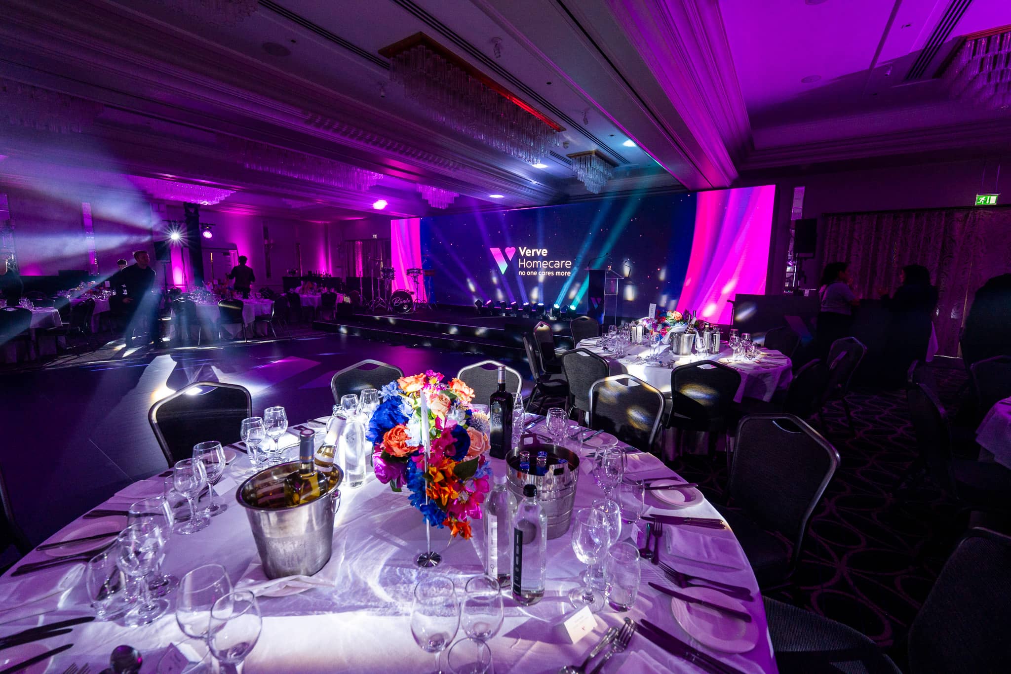 A floral centrepiece and table setting at Verve Awards Night, with purple uplighting and the LED wall and stage in the background at Sopwell House.