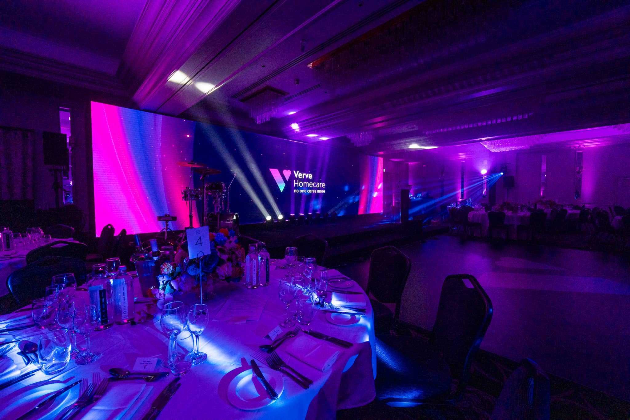 Round table place settings in purple light at Verve Awards Night, with the Verve Homecare LED wall and beam lighting beyond, St Alban Suite, Sopwell House.