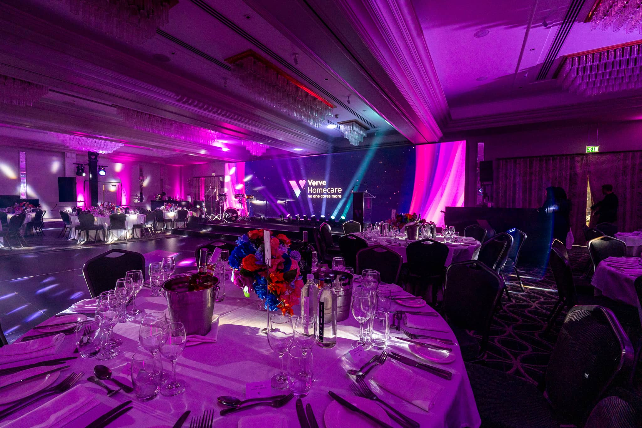 Dinner tables with floral centrepieces under purple uplighting, with the stage and LED wall across the St Alban Suite at Sopwell House.