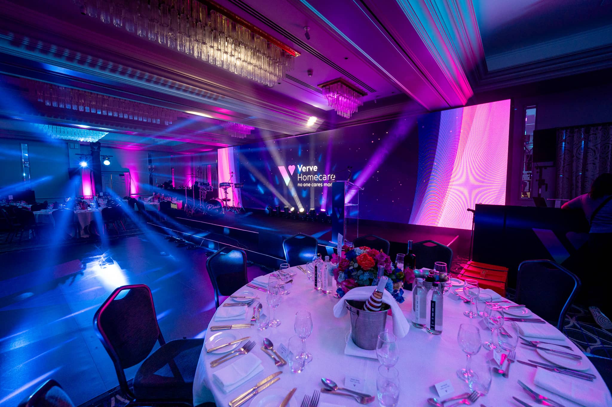 A round dinner table under blue and purple light, with beams crossing towards the Verve Homecare LED wall and stage at Sopwell House.
