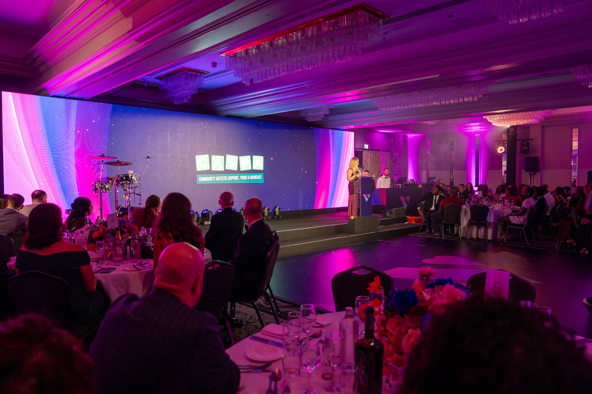 Guests watch a presentation beneath the chandeliers at Verve Awards Night, with the LED wall, band backline and magenta uplighting at Sopwell House.
