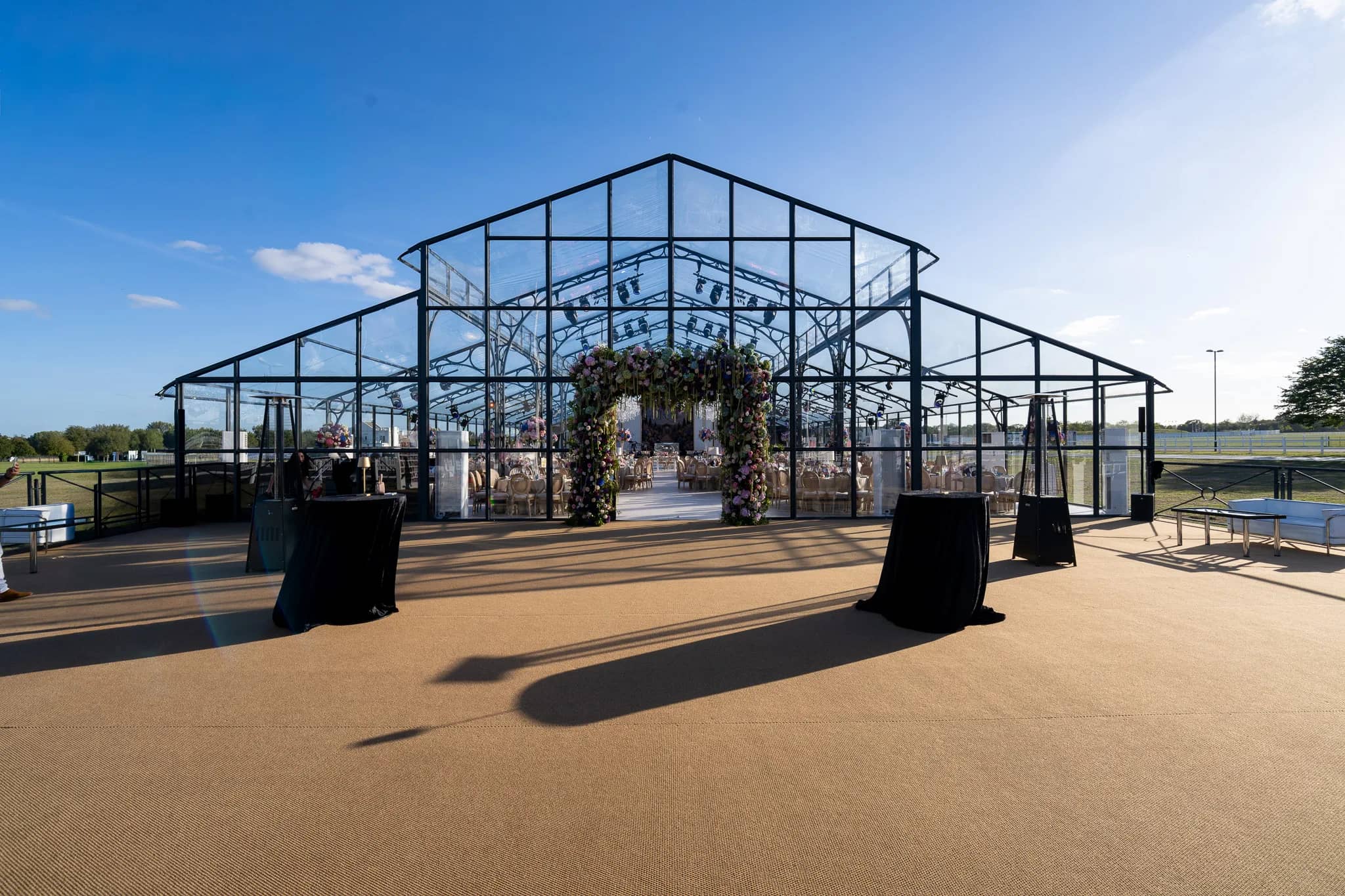 The front of a glass marquee with a floral arch at its door and poseur tables on a coir forecourt, Warwickshire, by Kudos AV