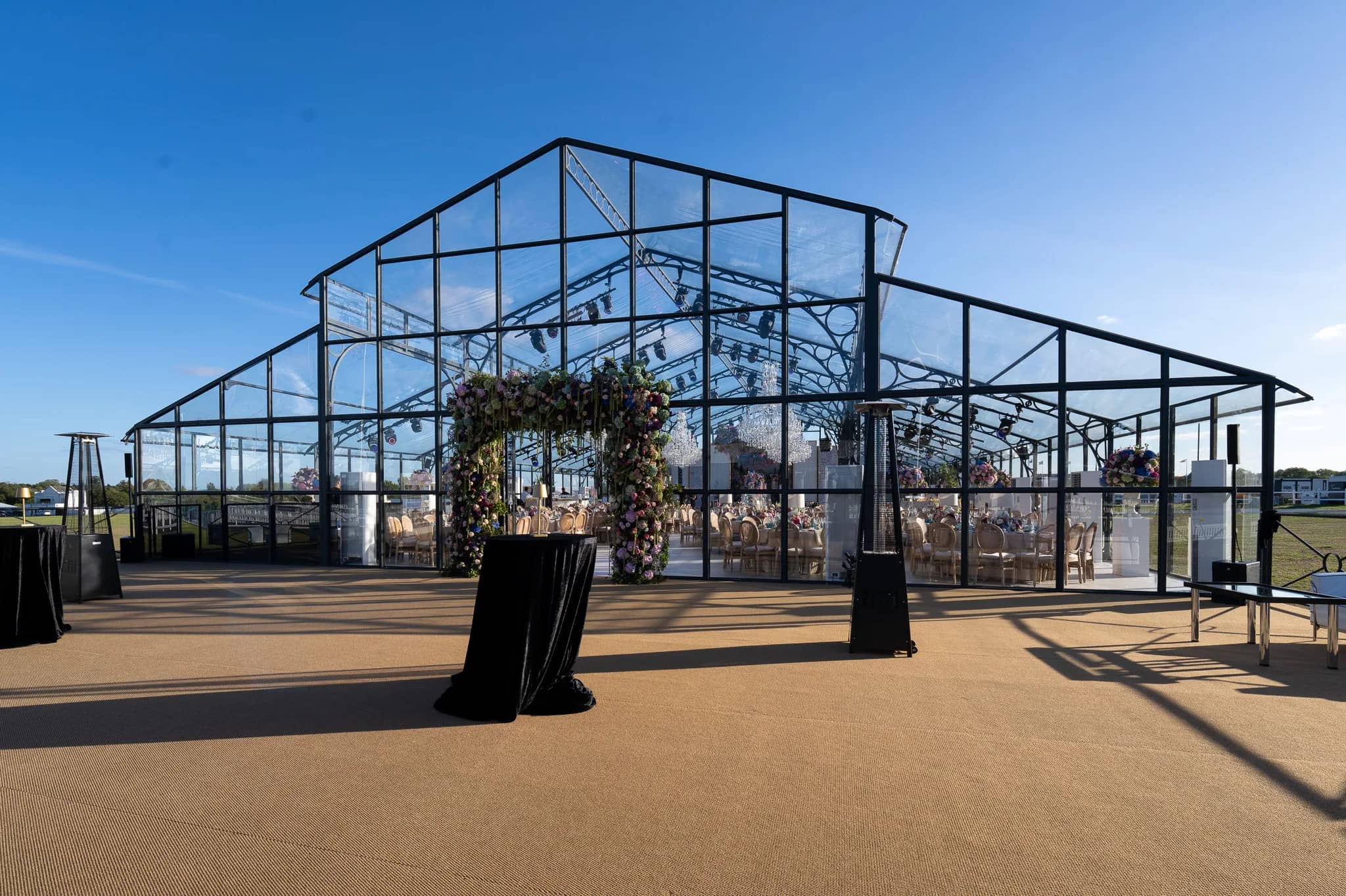 The floral arch and the forecourt of a glass marquee from the side, the tables and chandeliers visible through the glass, Warwickshire, by Kudos AV