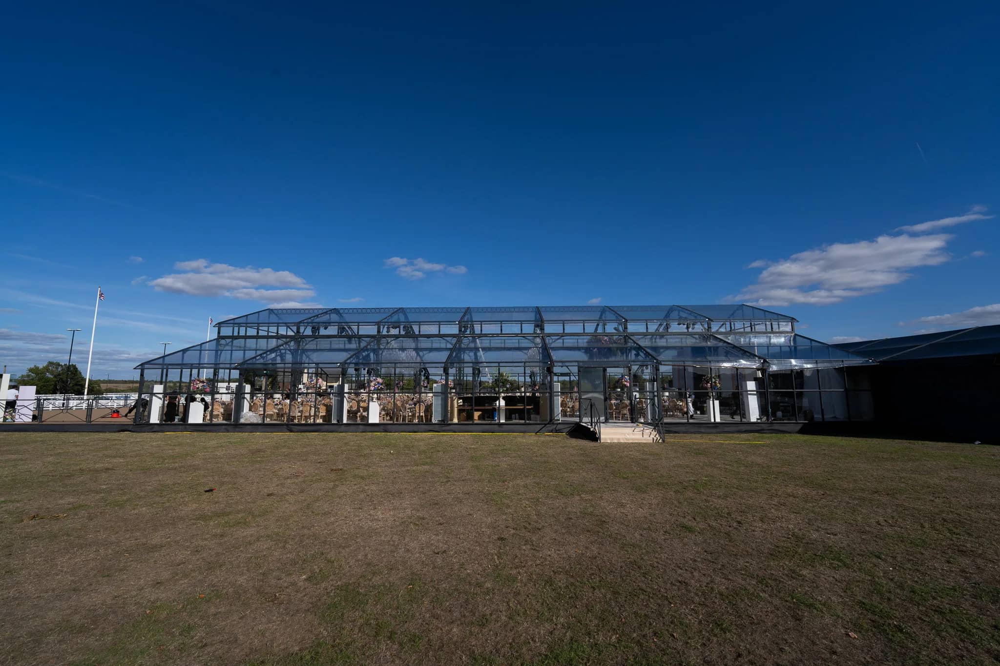 A 35 metre glass marquee by The English Marquee Company on the polo grounds under a blue sky, tables set inside, at Dallas Burston Polo Club, by Kudos AV