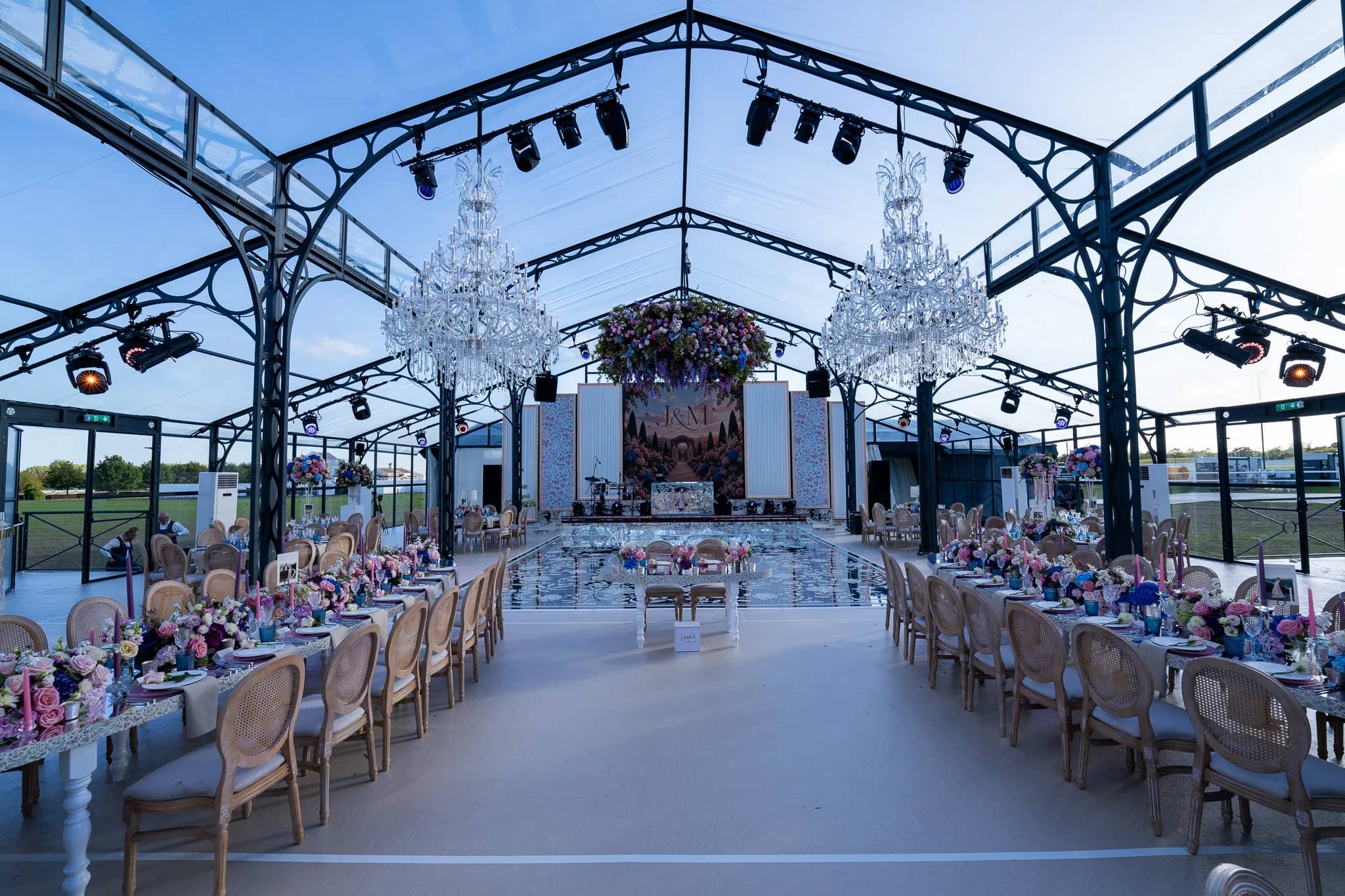 The room set by daylight: long tables of wildflowers, a printed mirror dancefloor reflecting the sky, a floral chandelier above it and the stage at the far end, glass marquee in Warwickshire, by Kudos AV
