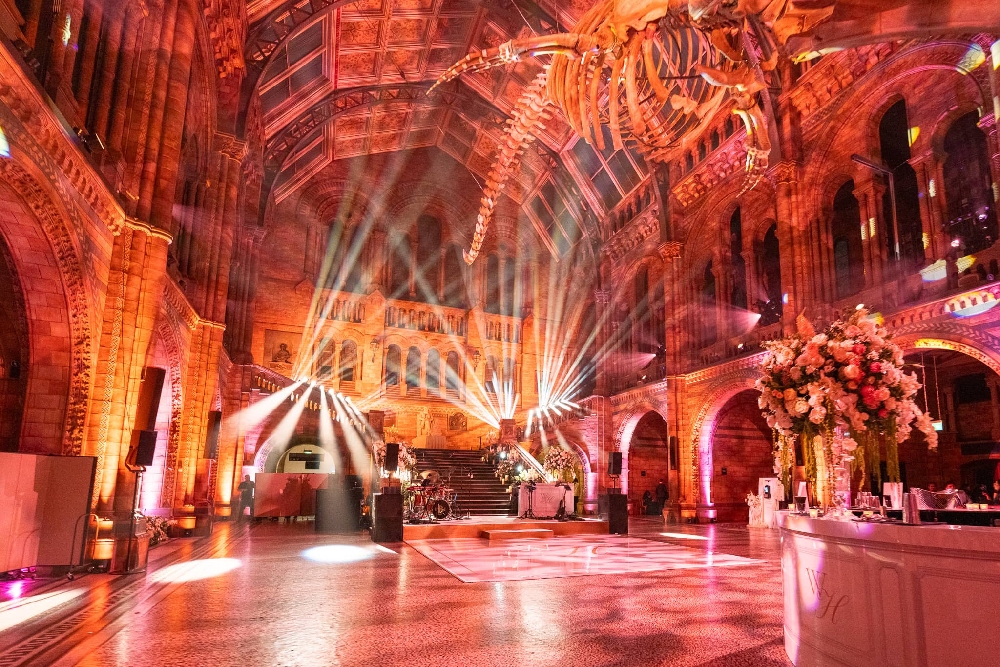 Crossing beams and red and amber uplighting through the full height of Hintze Hall, the party lighting at a Natural History Museum wedding by Kudos AV.
