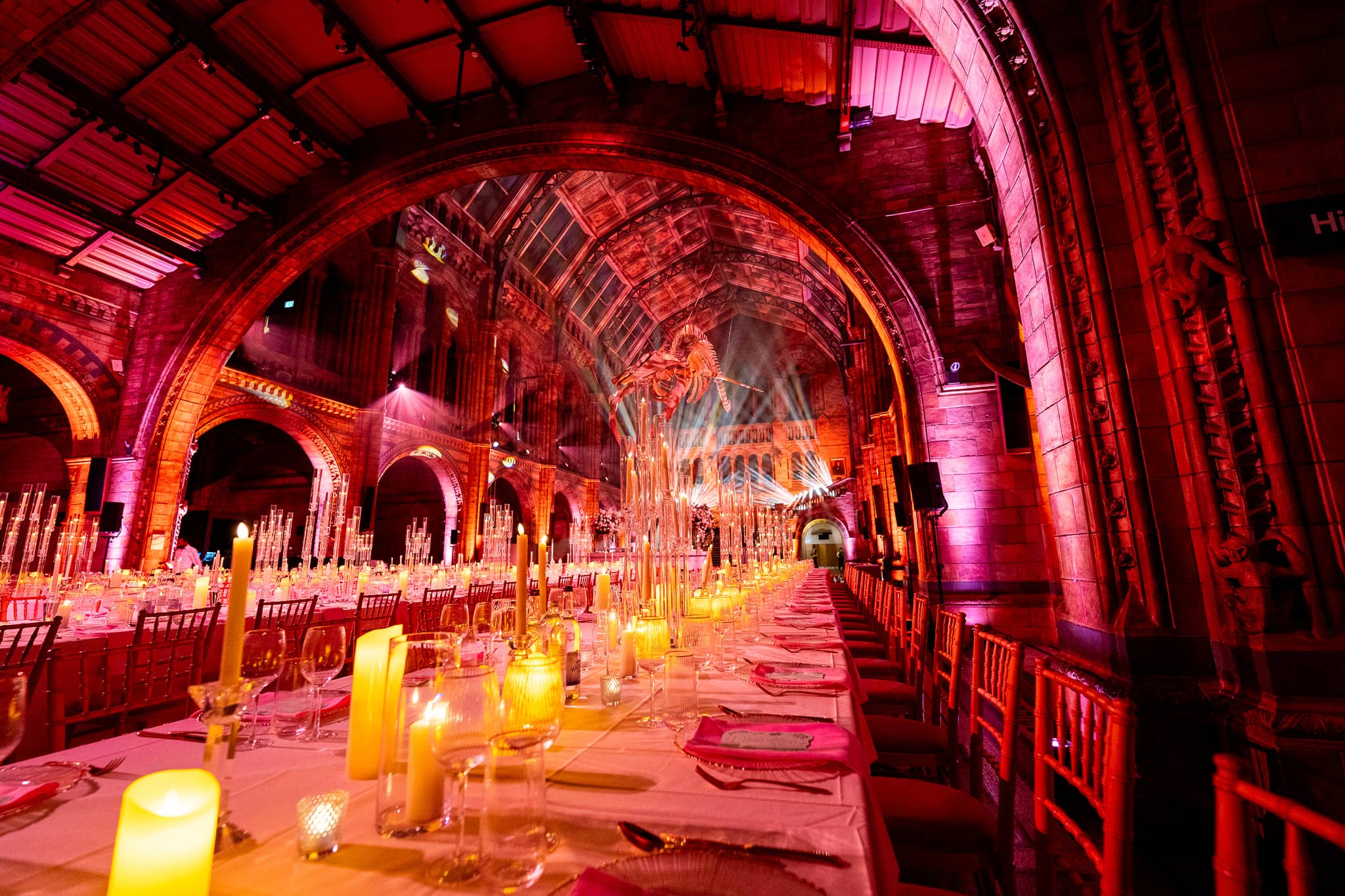 Red and pink uplighting across the vaulted ceiling of Hintze Hall above long banquet tables set with candles, a Natural History Museum wedding by Kudos AV.