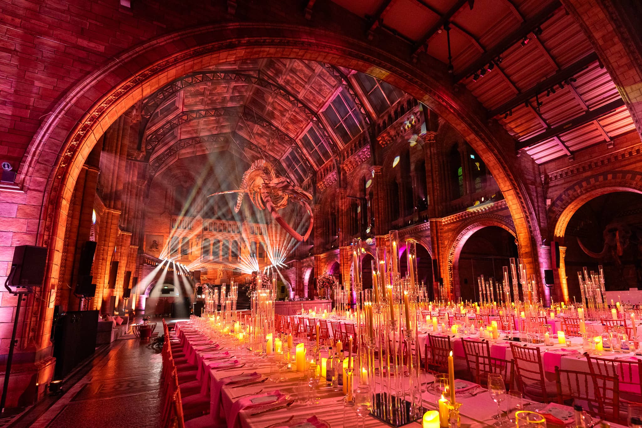 Candlelit banquet tables in Hintze Hall with gobo patterns and red and amber uplighting across the stonework, a Natural History Museum wedding by Kudos AV.
