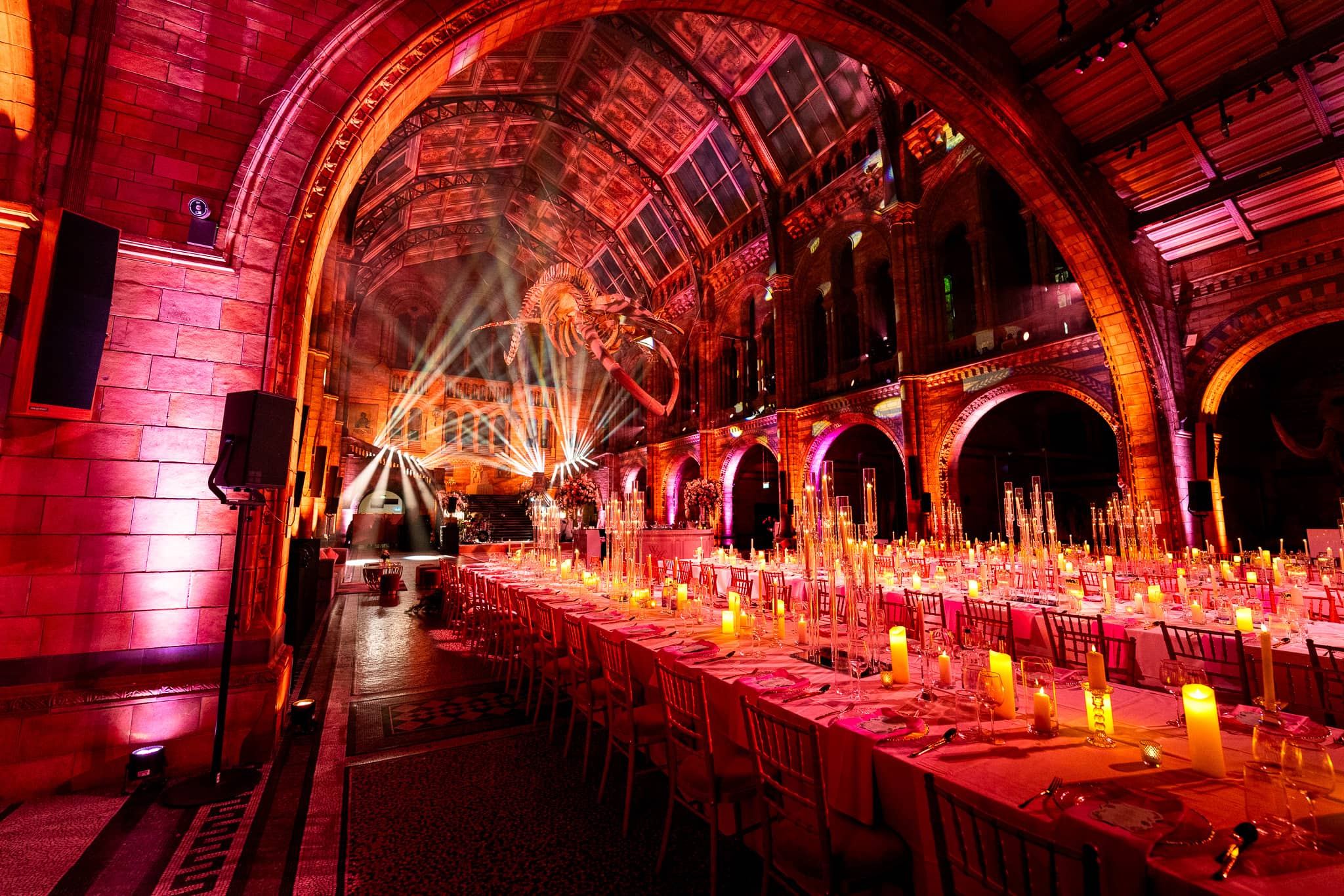 Long candlelit dinner tables down Hintze Hall under red uplighting on the arched ceiling, a 200 guest wedding at the Natural History Museum lit by Kudos AV.