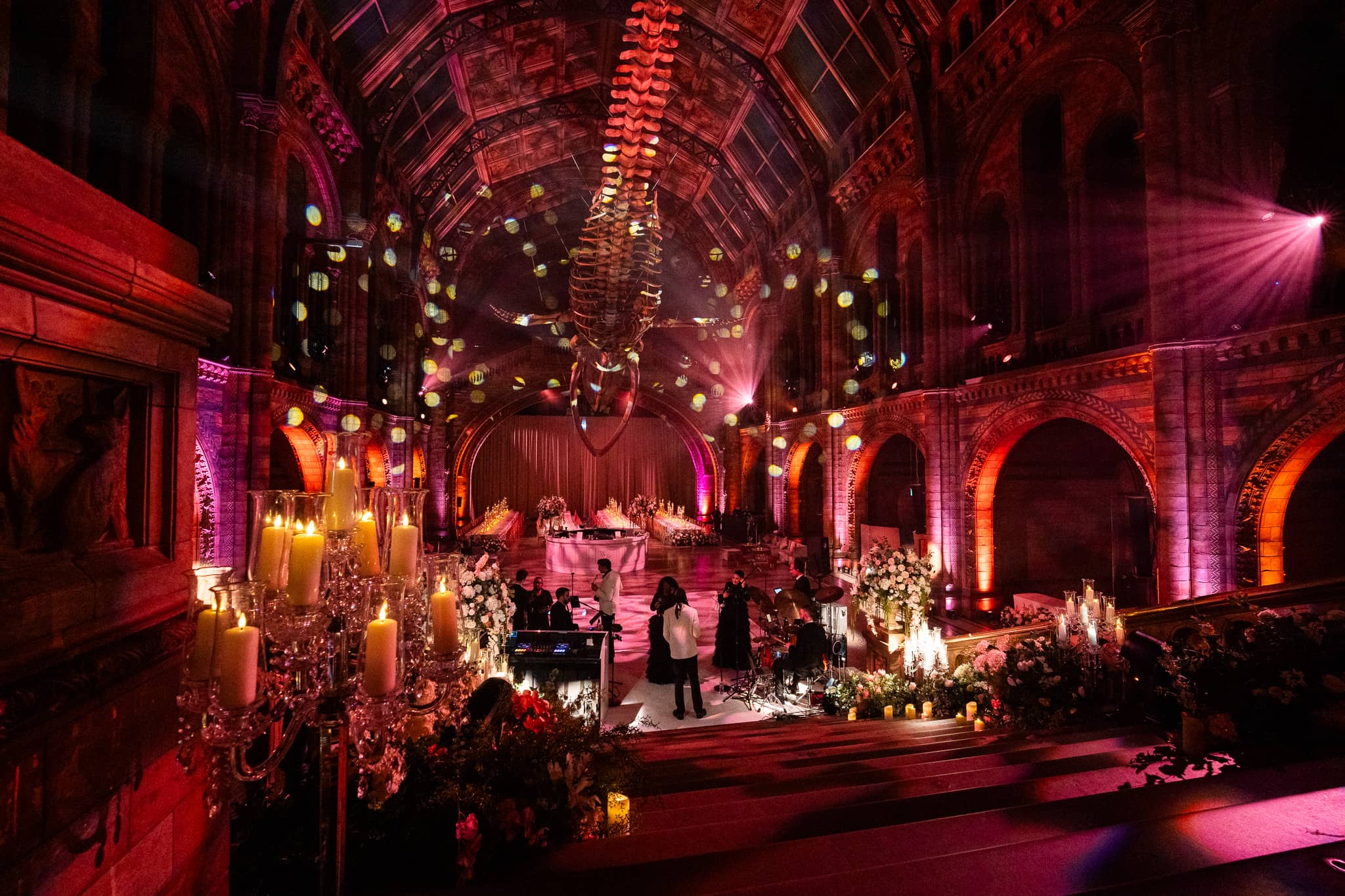 Red and pink uplighting over candles, florals and hanging lanterns in Hintze Hall, the front of the hall lit for the party, at a Natural History Museum wedding by Kudos AV.