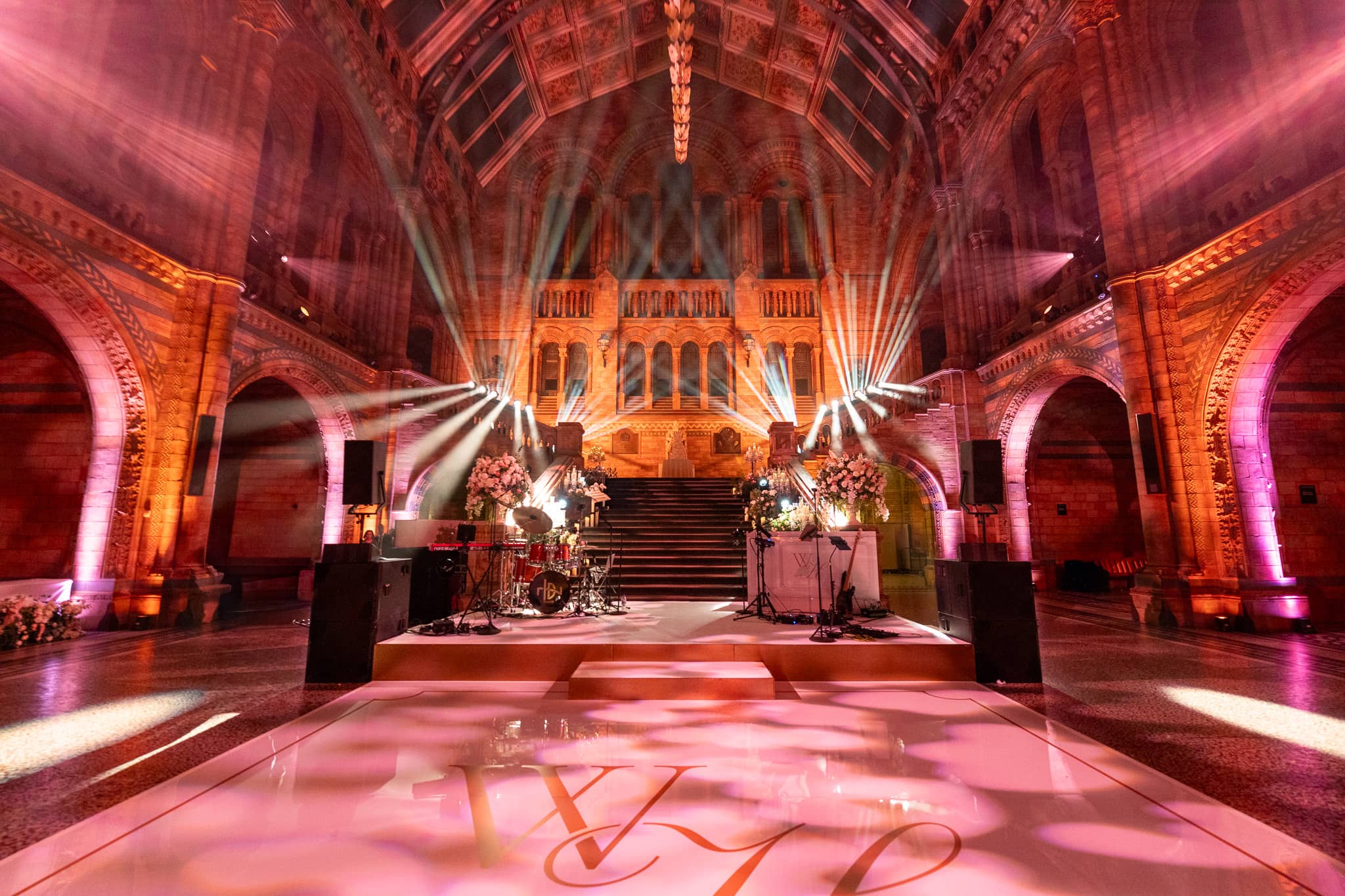The printed dancefloor at the centre of Hintze Hall under pink and amber uplighting, florals alongside, at a Natural History Museum wedding by Kudos AV.