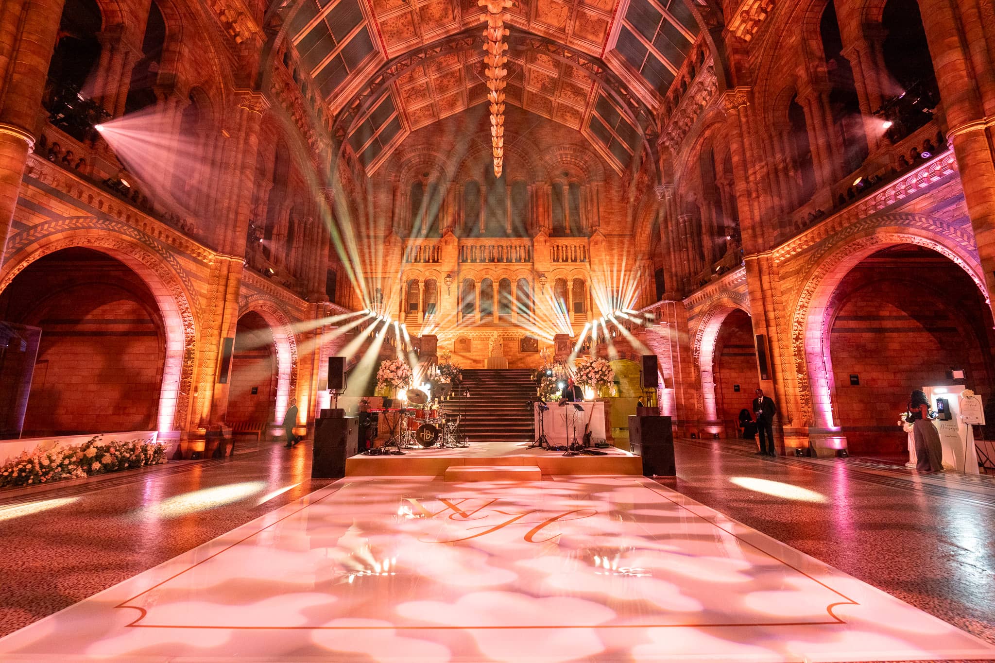 Beams and pink uplighting filling Hintze Hall for the party, florals at the centre of the hall, at a Natural History Museum wedding by Kudos AV.