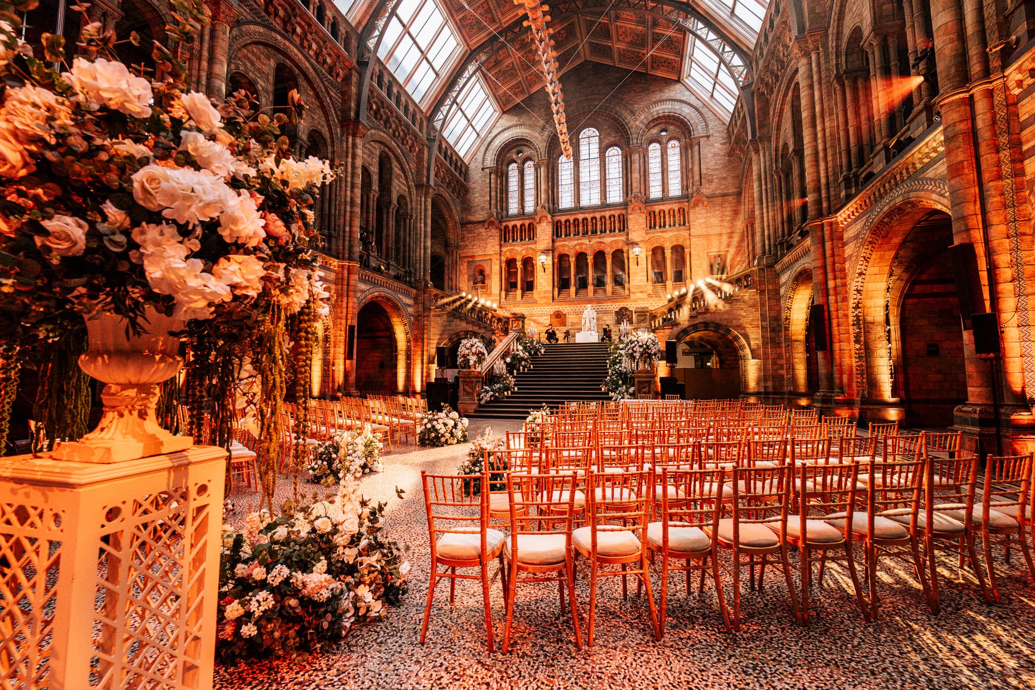 Rows of ceremony chairs facing the floral installation in Hintze Hall, warm uplighting on the stonework, at a Natural History Museum wedding by Kudos AV.