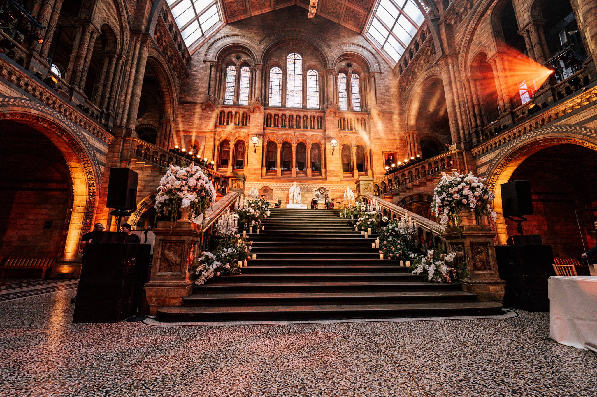The main staircase in Hintze Hall uplit in amber with white florals on the steps, set for a wedding at the Natural History Museum by Kudos AV.