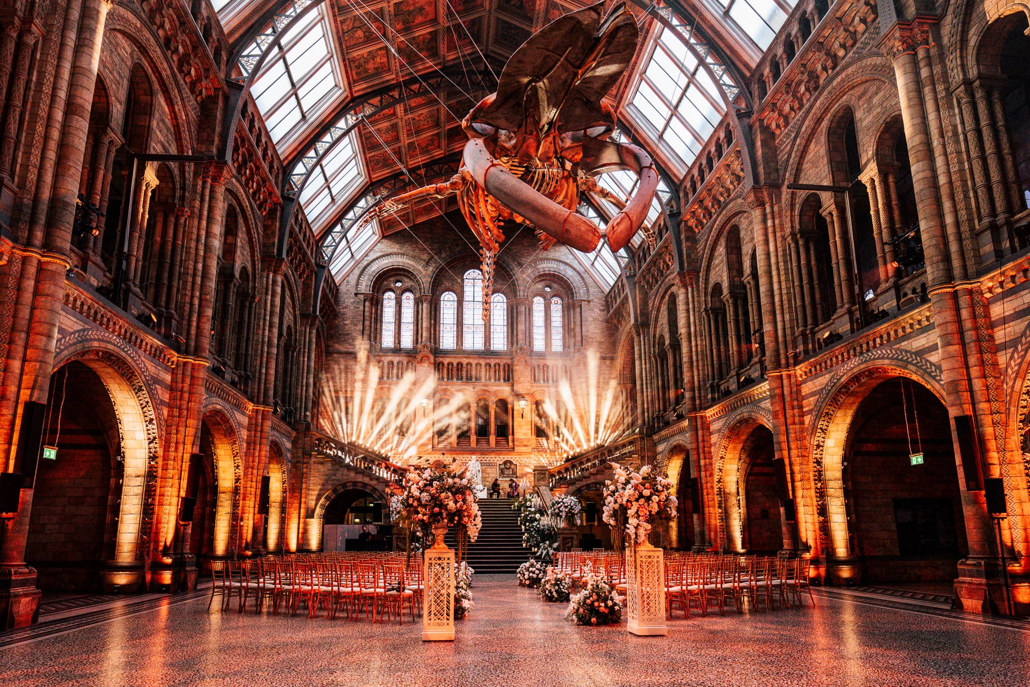 The ceremony aisle in Hintze Hall under red lighting, Hope the blue whale suspended above the seating and florals, lit by Kudos AV.