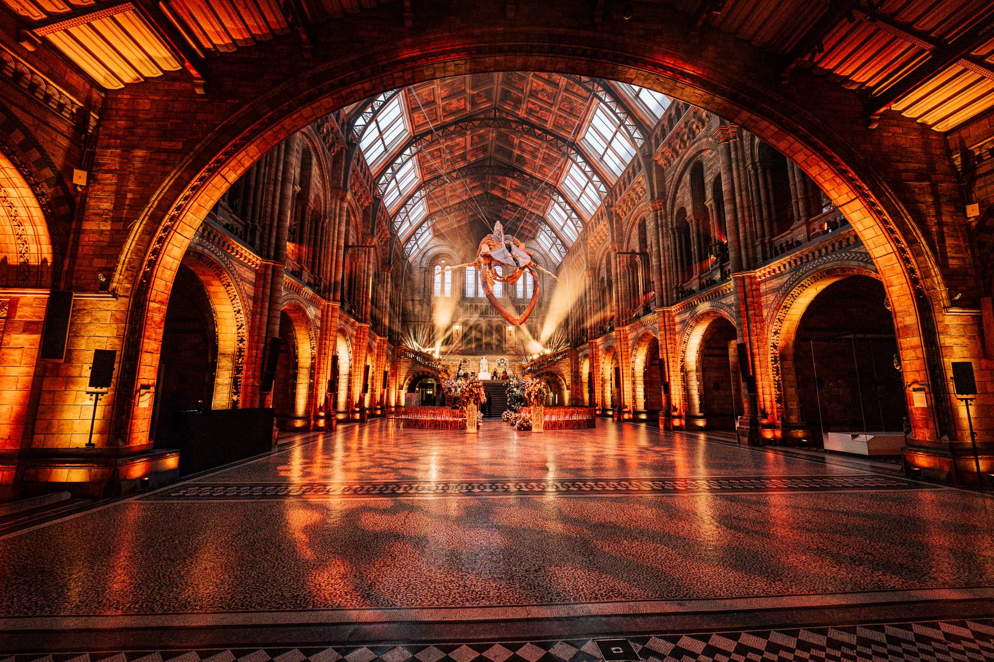 The terracotta arches and vaulted ceiling of Hintze Hall washed in warm amber uplighting before a wedding at the Natural History Museum, lit by Kudos AV.
