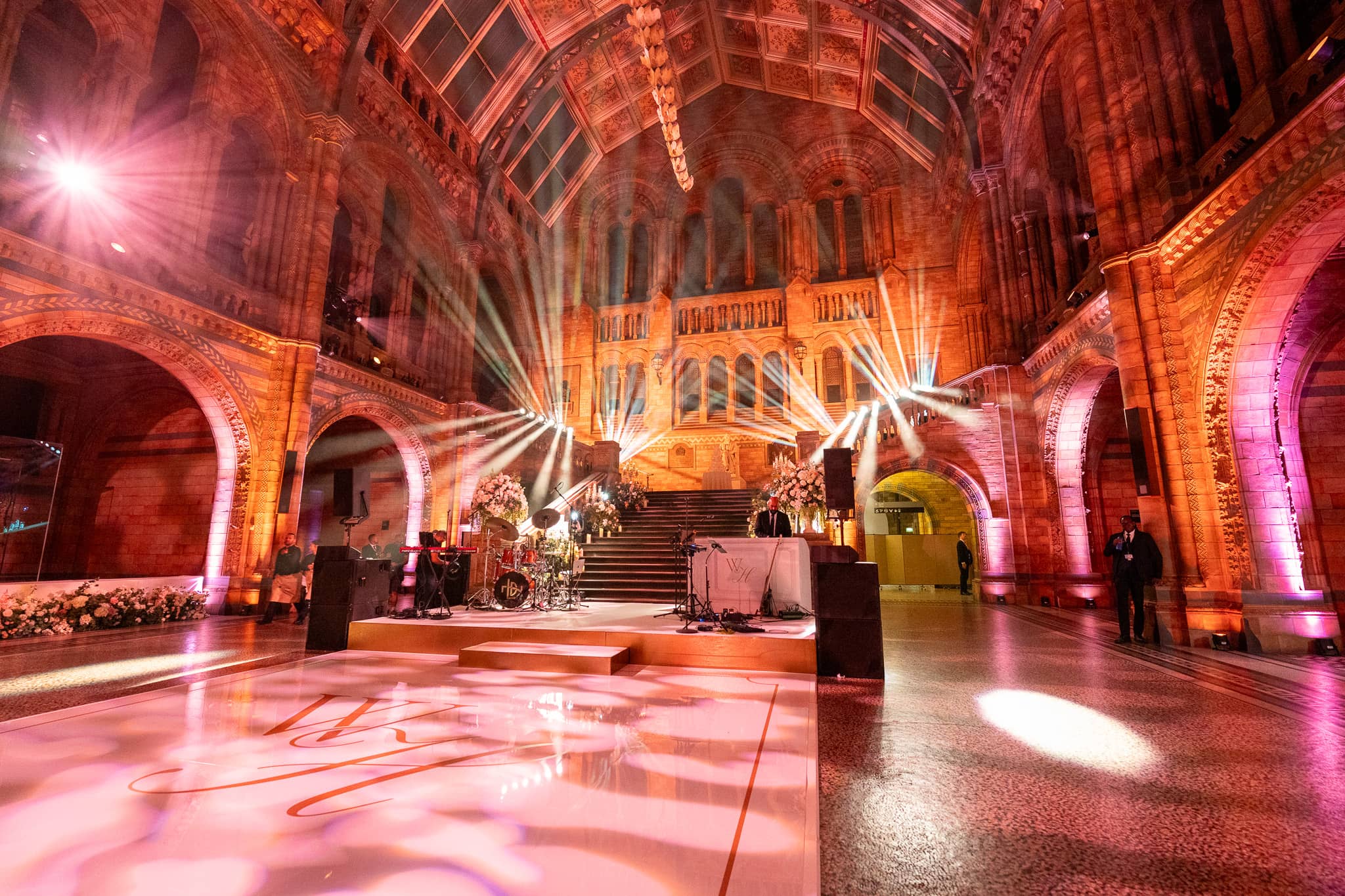 Pink and amber uplighting with beams through the vault of Hintze Hall, florals at the centre of the hall, a Natural History Museum wedding lit by Kudos AV.