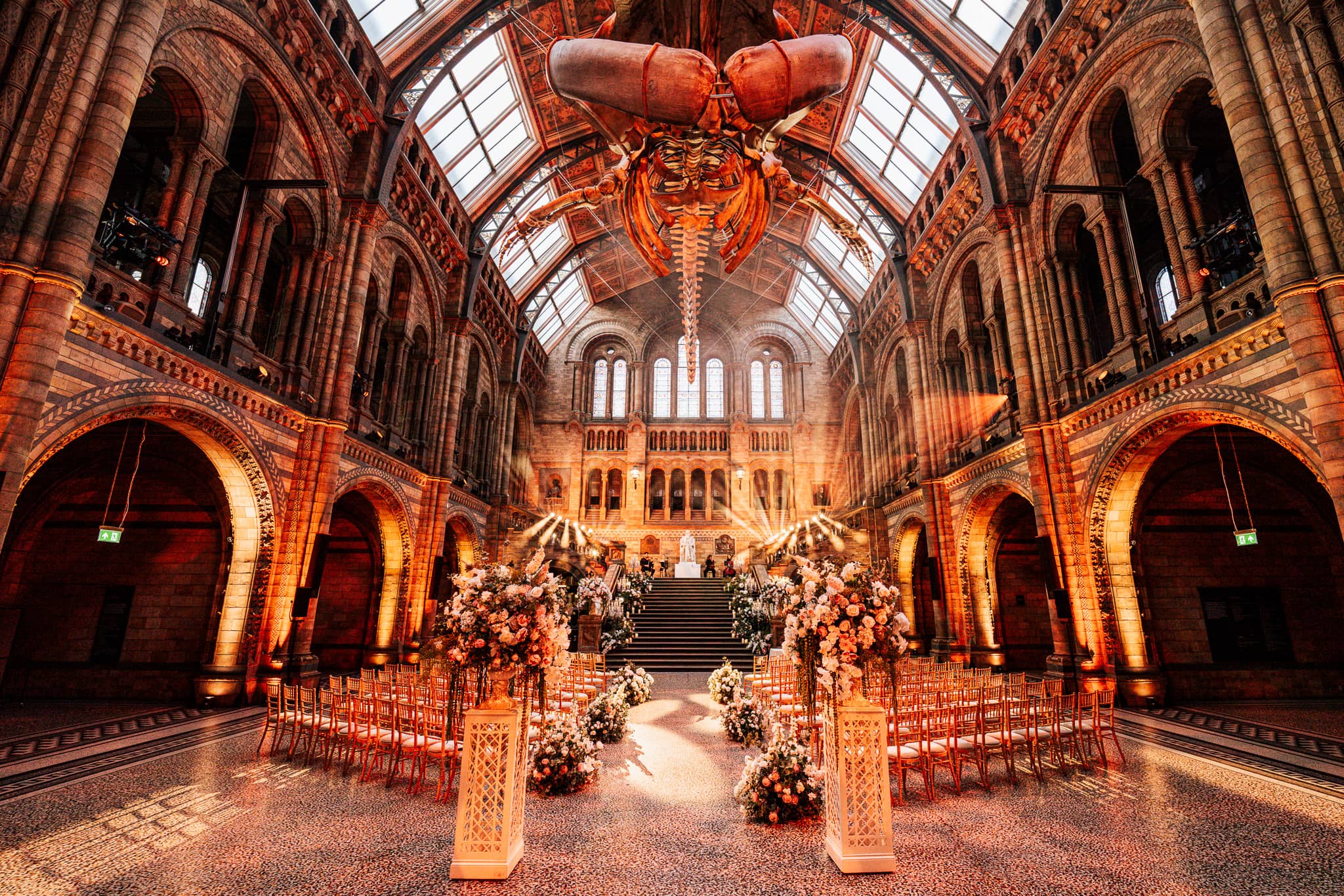 Ceremony florals beneath the arches and glass roof of Hintze Hall with Hope the blue whale above, at a Natural History Museum wedding lit by Kudos AV.