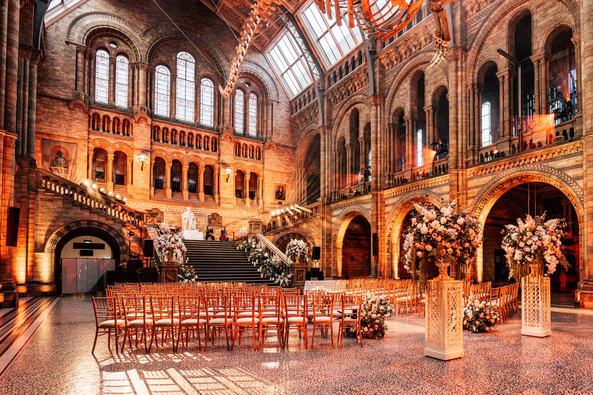 Ceremony seating and florals in Hintze Hall under warm uplighting, the arches and glass roof of the Natural History Museum above, by Kudos AV.