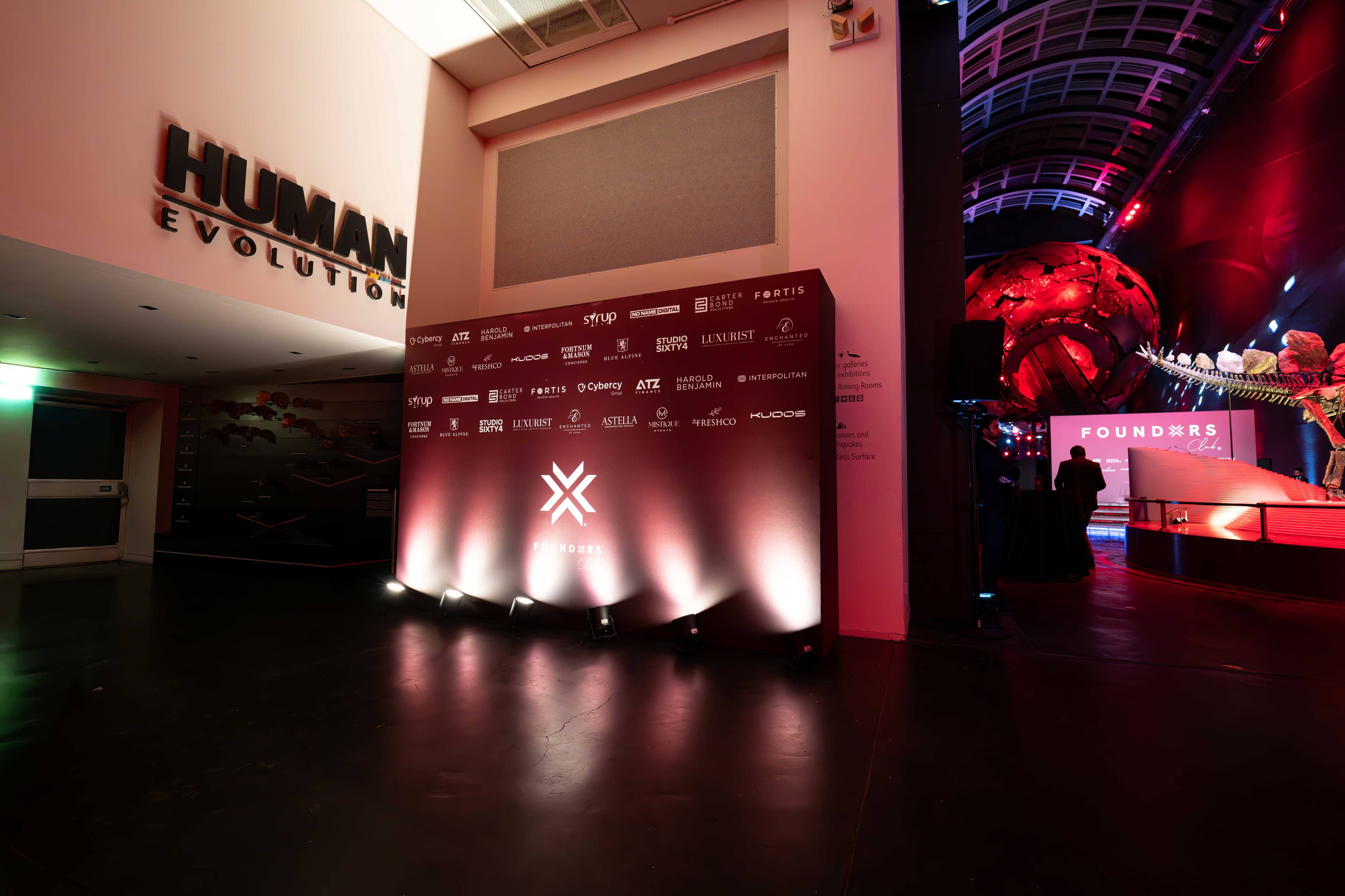 The FOUNDXRS partner wall under red uplighting beside the museum’s gallery signage, FOUNDXRS Club black tie party at the Natural History Museum, by Kudos AV.