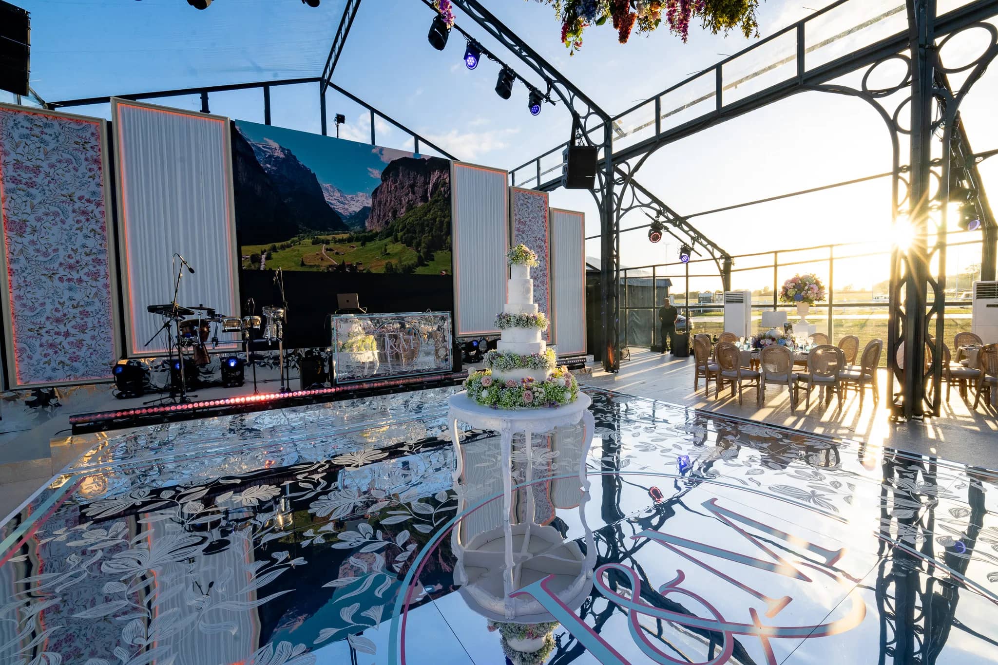 An alpine valley on the central LED screen at sunset, the mirror-printed DJ booth and a drum kit on stage and the cake on the mirror floor, glass marquee at Dallas Burston, by Kudos AV
