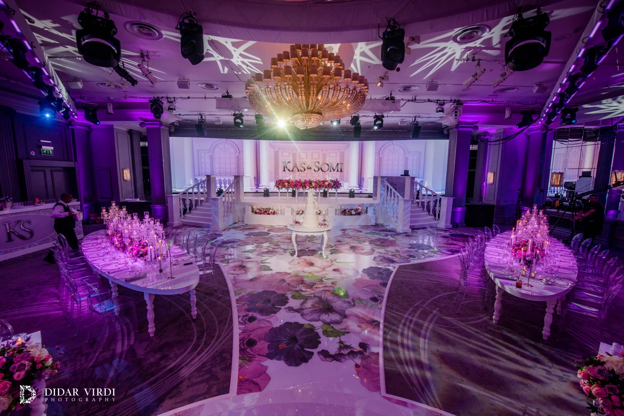 Moving lights and patterned ceiling washes above the white wedding stage, floral walkway and dining tables in the Grosvenor House Ballroom.