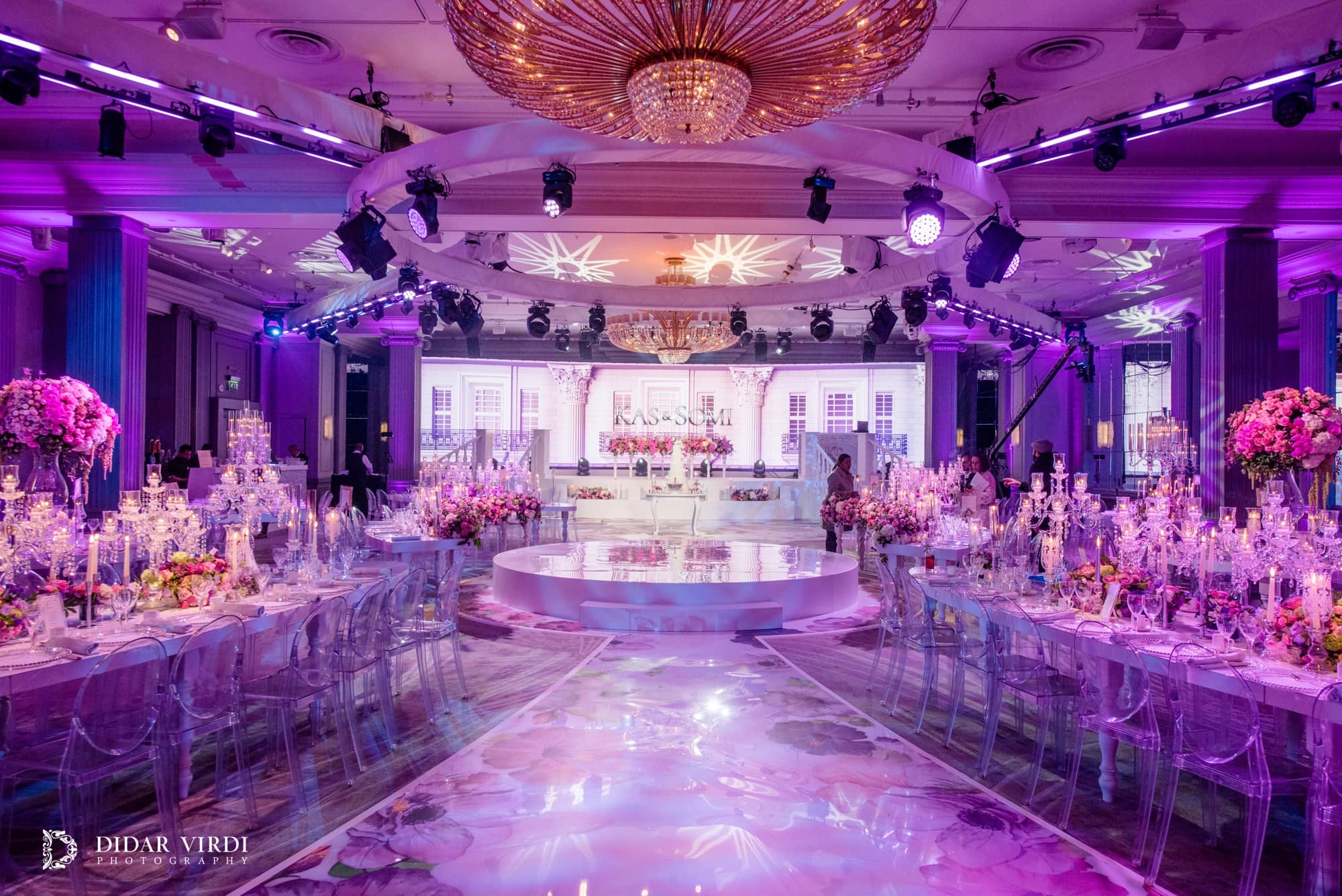 View along the floral wedding walkway towards the white stage, with oval tables and overhead lighting in the Grosvenor House Ballroom.