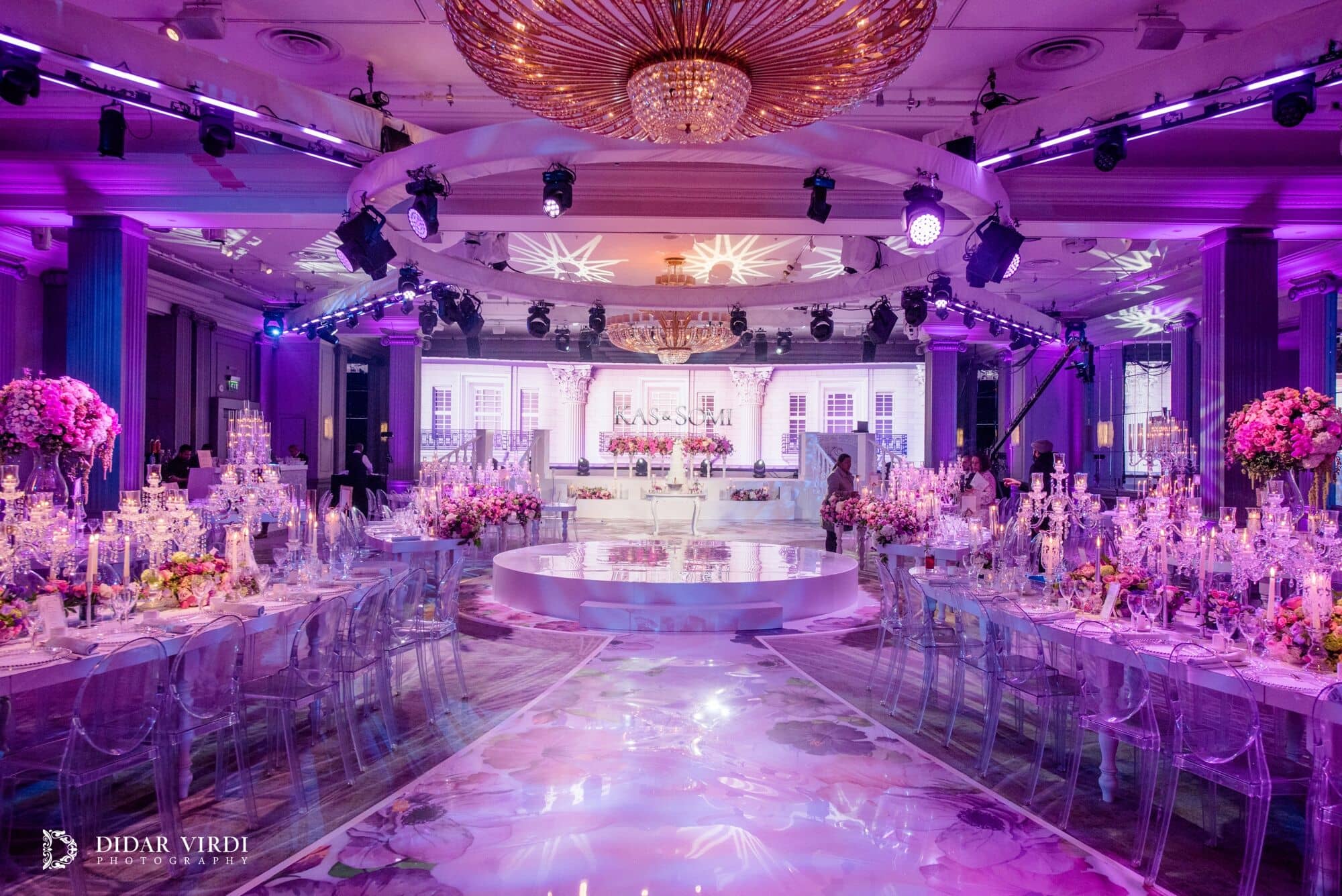 Purple colour washes light the tables and floral walkway leading to the white wedding set in the Grosvenor House Ballroom.