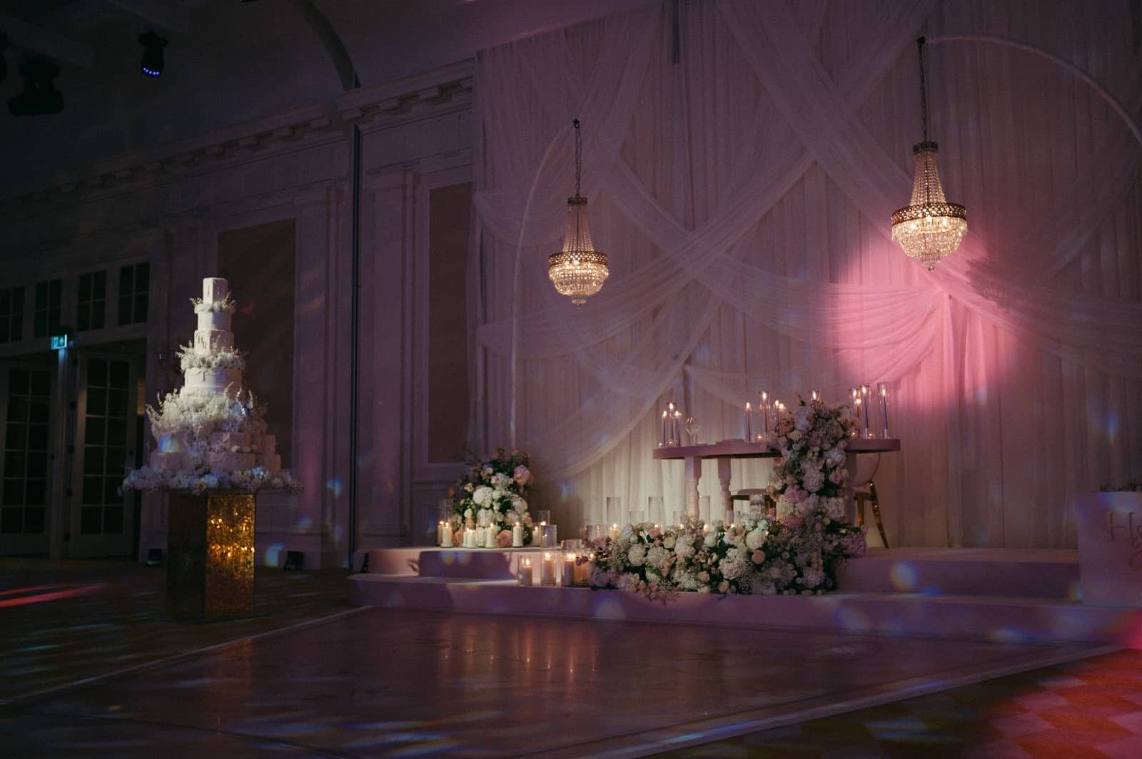 Head table stage backed by pink-lit drapes and pendant lights at Raffles London, with a tiered cake beside the stage.