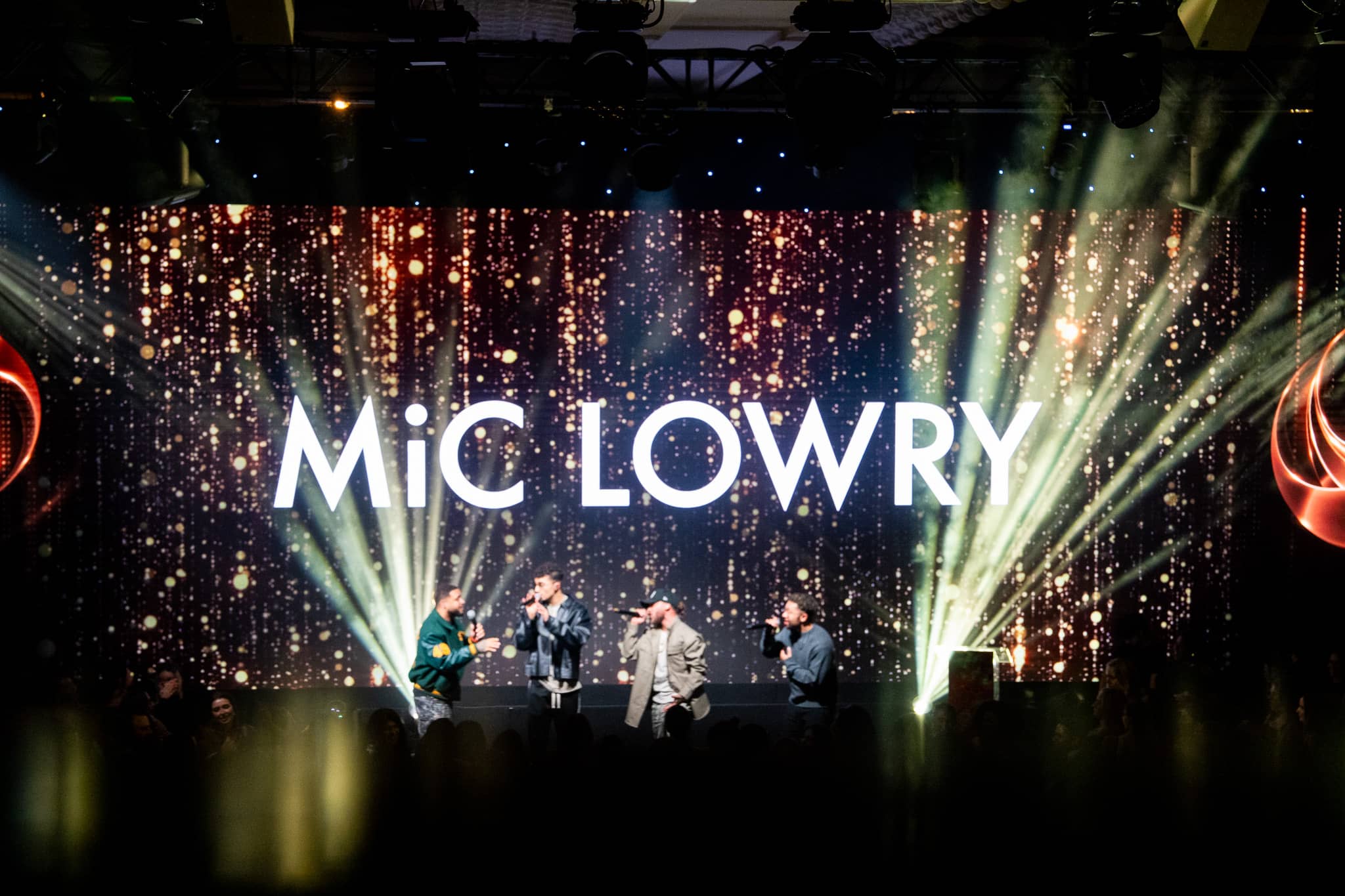 MiC LOWRY under green and amber stage lighting with the LED backdrop lit behind, Dental Beauty Partners Awards 2025 in the Great Room at Grosvenor House London, by Kudos AV.