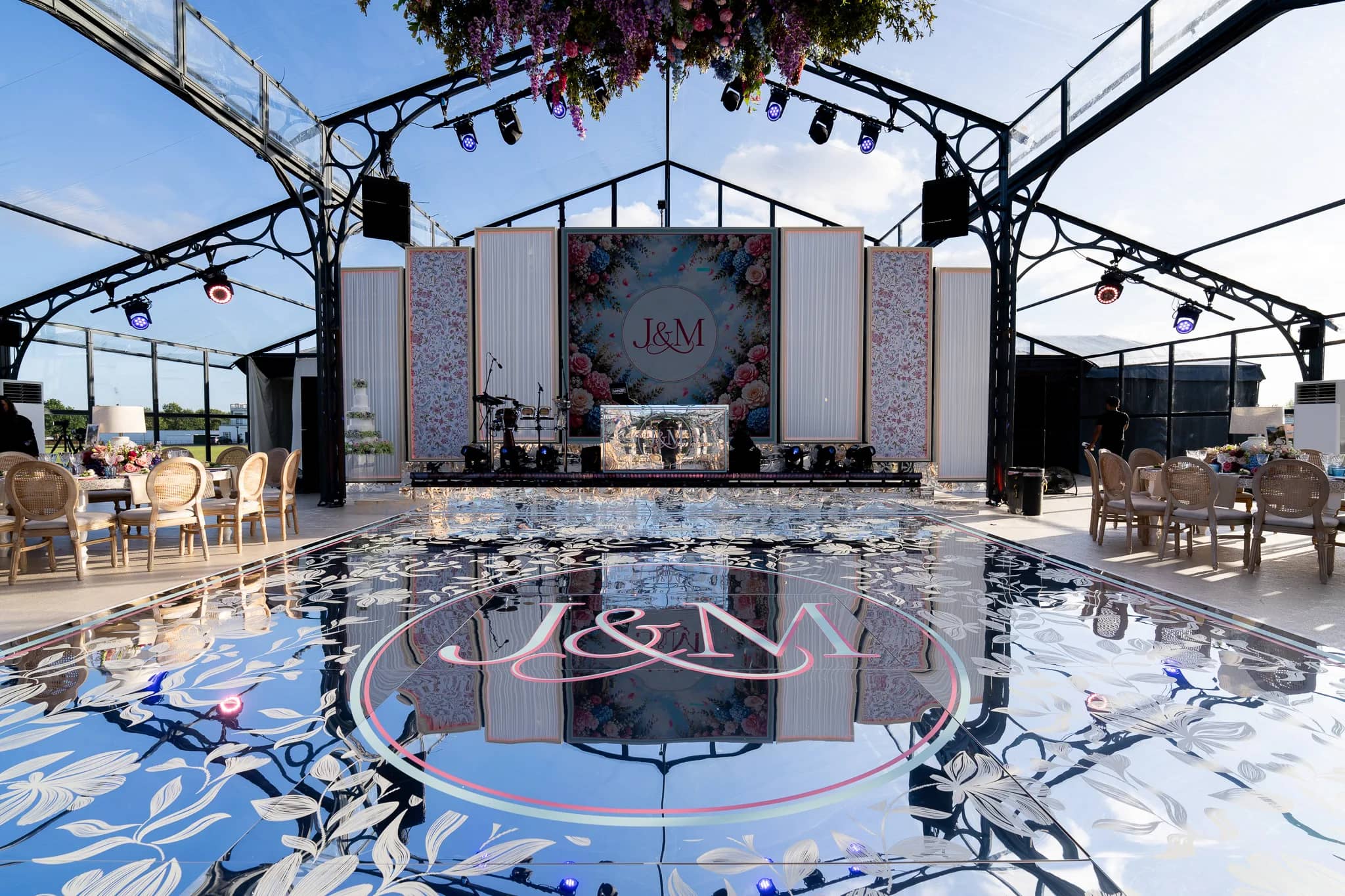 The stage by day: a central LED screen with a floral monogram, LED-edged drape panels either side, a mirror-printed DJ booth and a drum kit, and a printed mirror dancefloor with the monogram in front, glass marquee at Dallas Burston, by Kudos AV