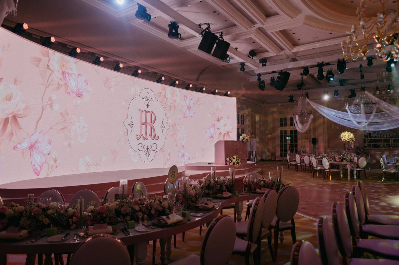 A wide LED screen displaying a pink floral monogram beneath the lighting rig, with dining tables in the foreground at Raffles London.