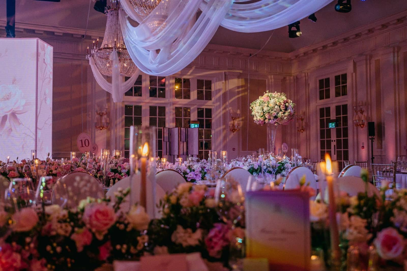 A floral table centrepiece and candles beneath draped chandeliers, with pink lighting and part of the LED screen behind at Raffles London.