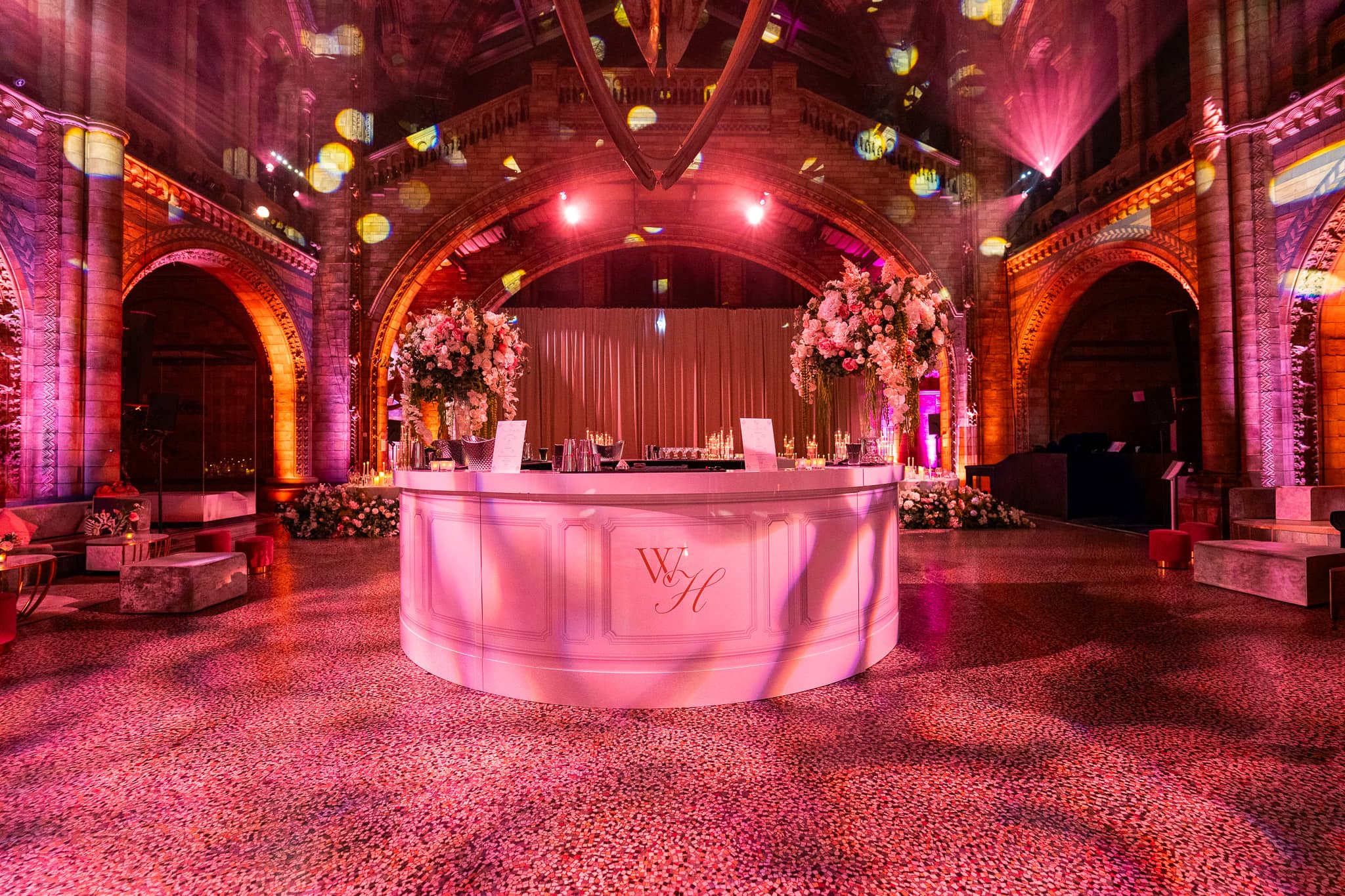 The printed circle bar glowing pink with the couple’s monogram and a floral arch behind, in Hintze Hall at the Natural History Museum, by Kudos AV.