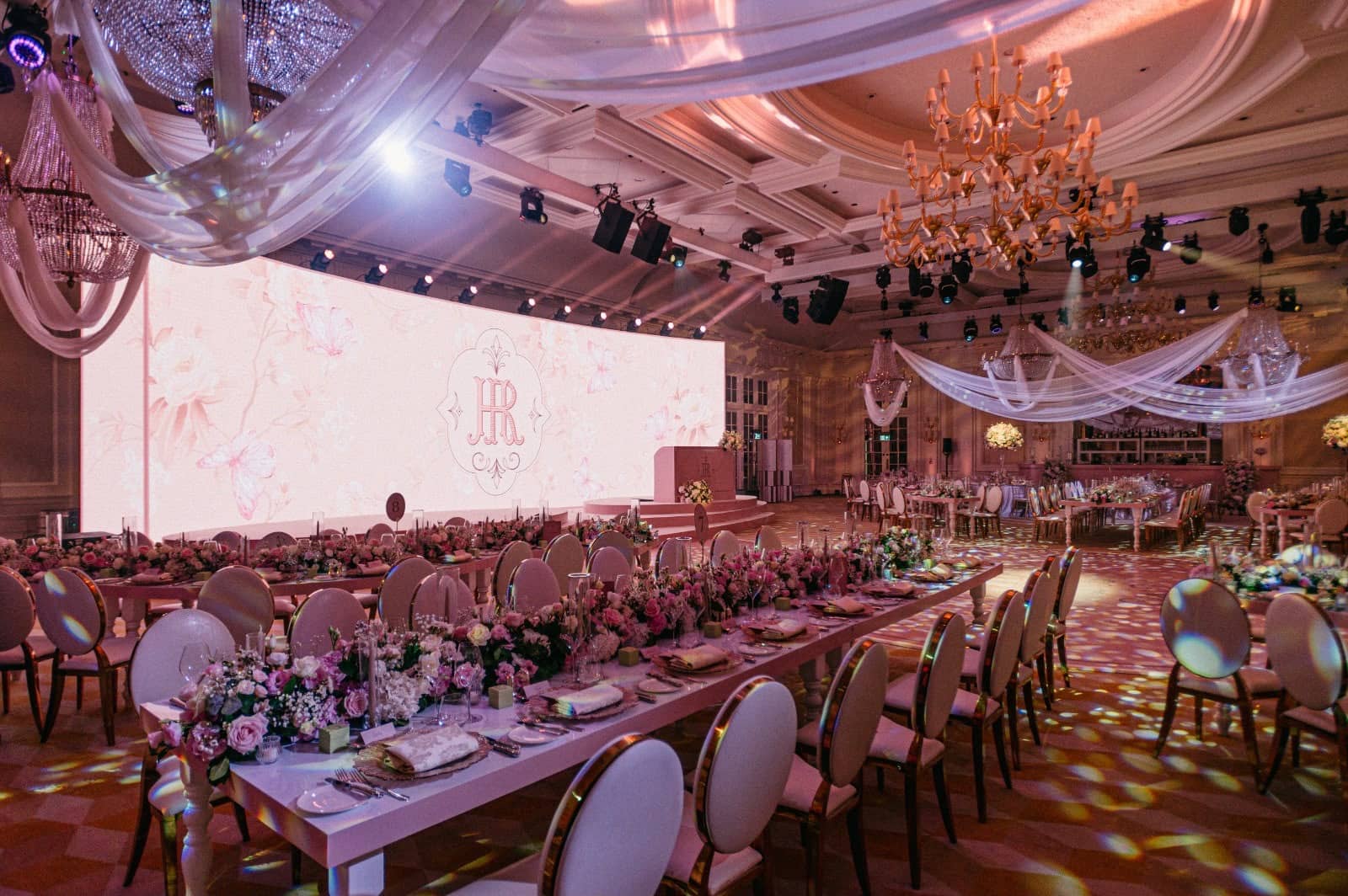 Long dining tables face a wide pink floral LED screen beneath crystal chandeliers, ceiling drapes and stage lighting at Raffles London.