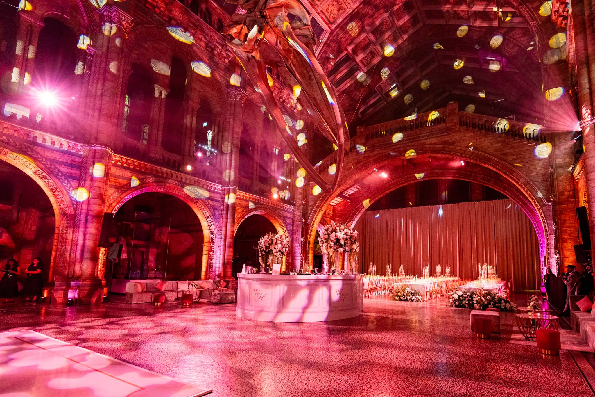 The circle bar in Hintze Hall under pink and magenta uplighting on the arches, florals around it, at a Natural History Museum wedding by Kudos AV.