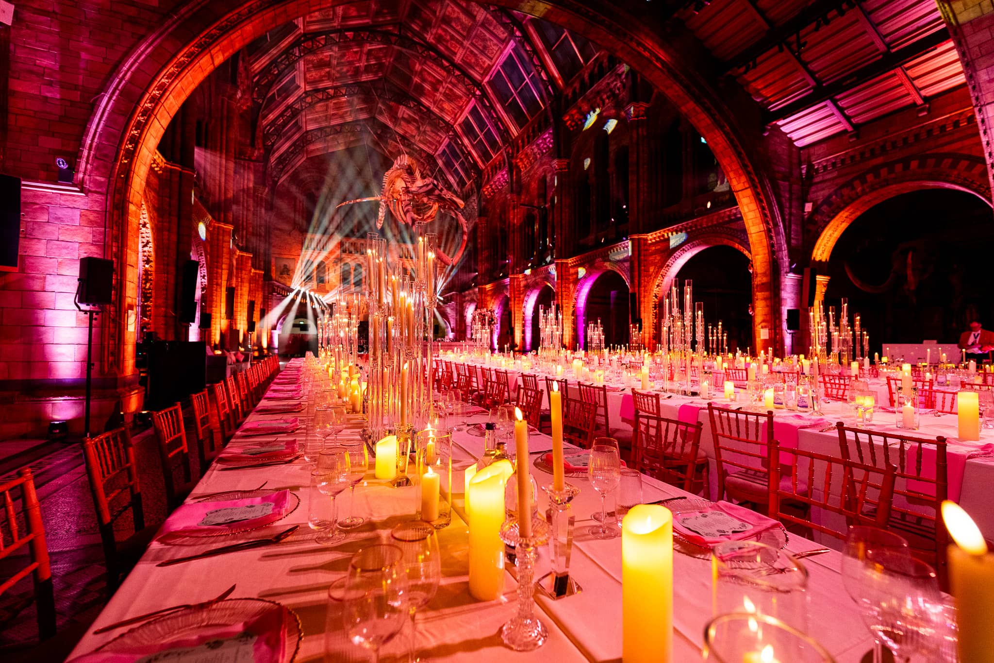 Long dinner tables with candlelit centrepieces in Hintze Hall under pink uplighting, the ornate arches above, at a Natural History Museum wedding by Kudos AV.