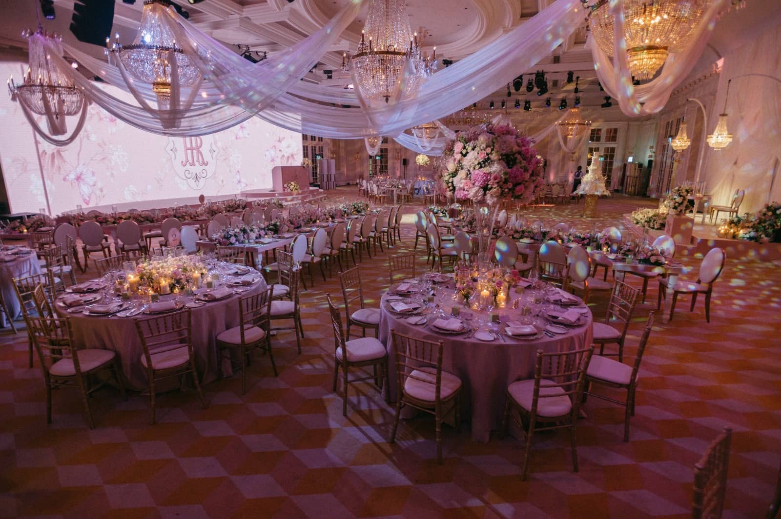 Round guest tables and floral arrangements beneath draped crystal chandeliers, with a pink LED backdrop along the ballroom at Raffles London.