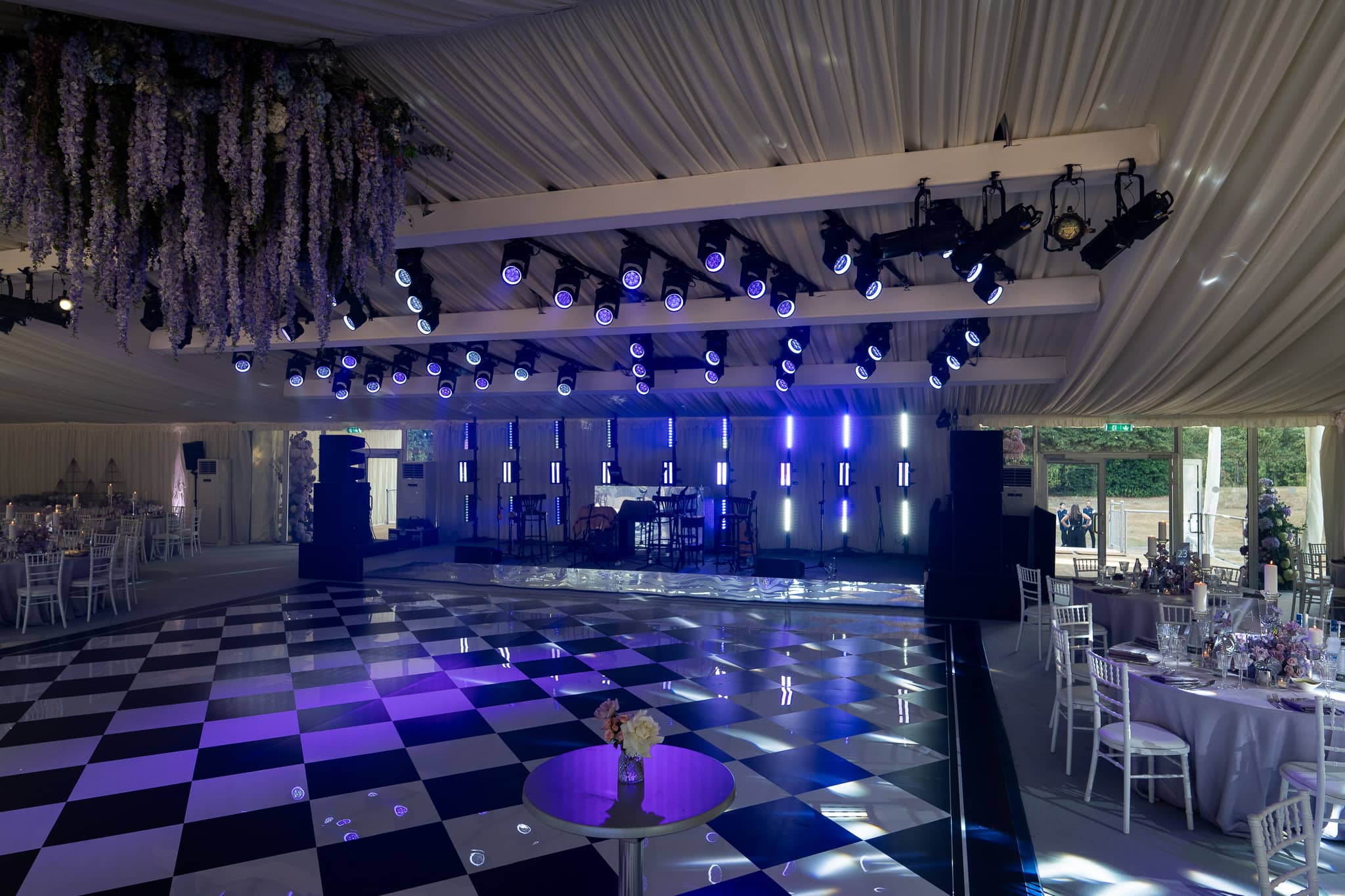 The lighting rig, a stage backdrop in blue and a lit chequerboard mirror dancefloor with florals hanging above, luxury marquee at Pennyhill Park, Surrey, by Kudos AV