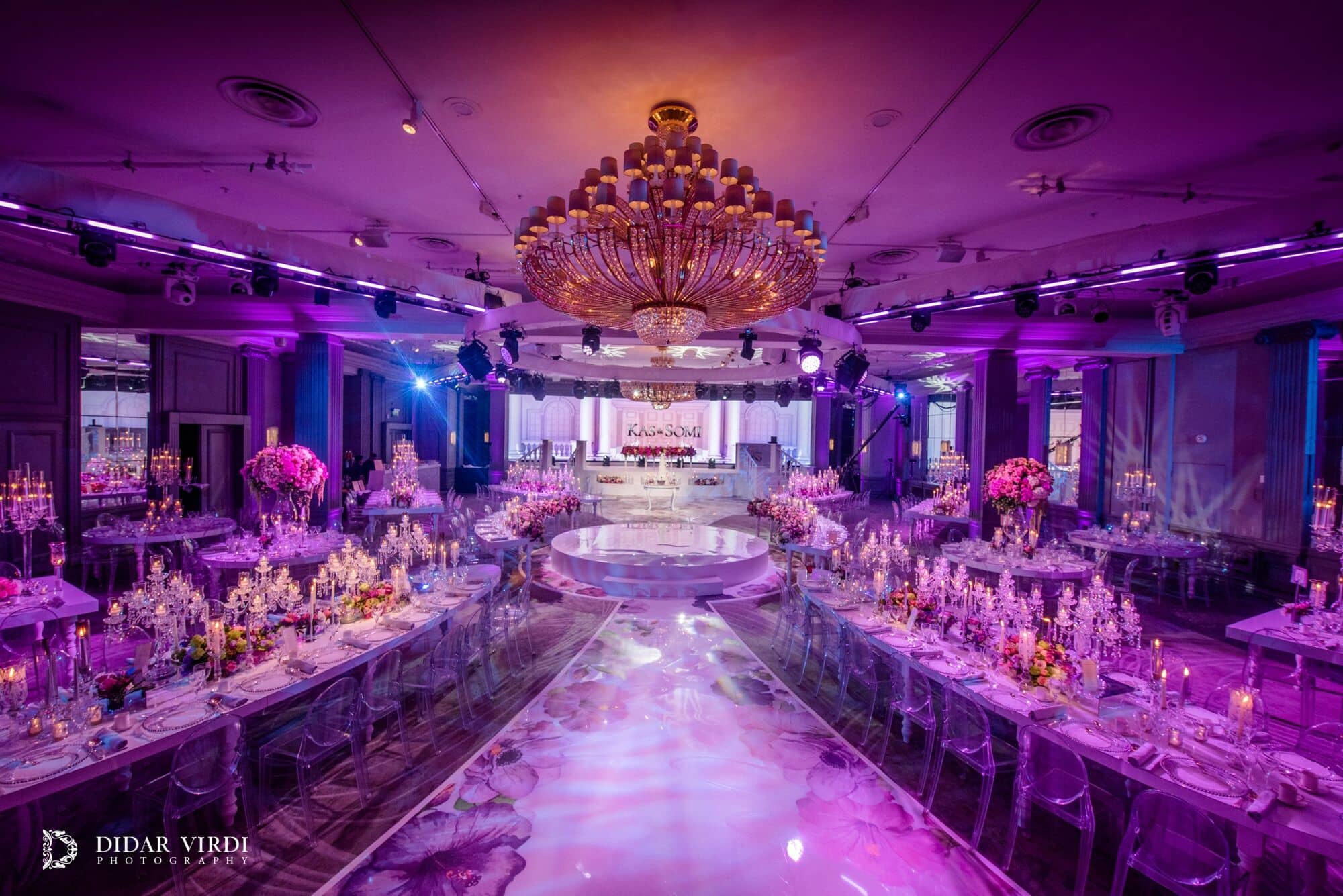 Wide view of the Grosvenor House Ballroom wedding, with chandeliers, purple lighting, curved tables and a floral walkway to the stage.
