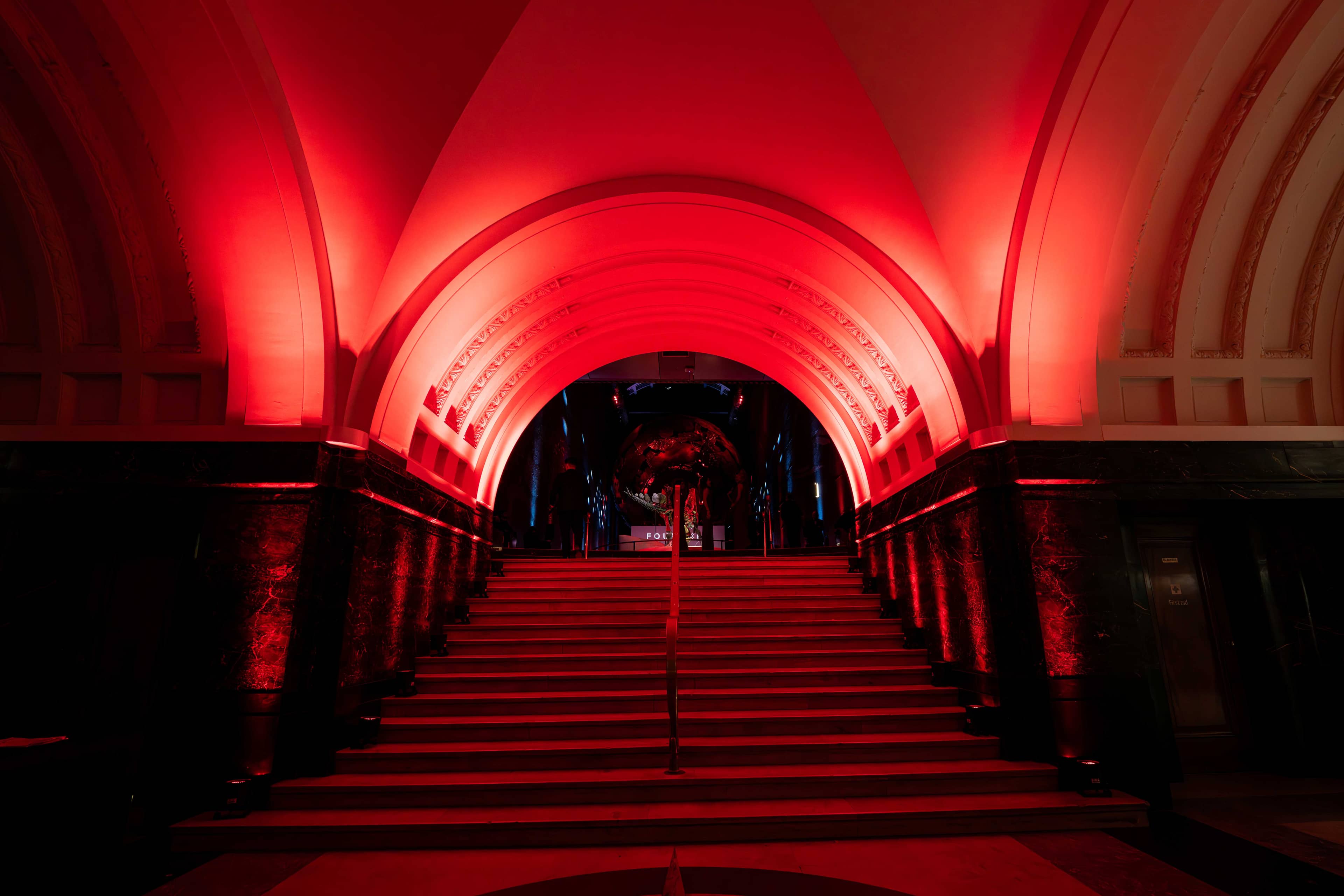 The museum’s stone arches and staircase lit red for the FOUNDXRS Club black tie party at the Natural History Museum, by Kudos AV.
