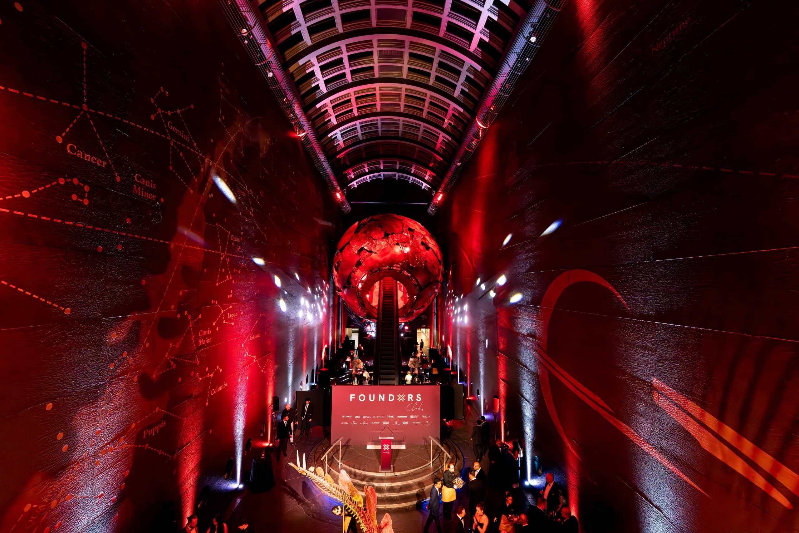 Earth Hall lit red for the FOUNDXRS Club black tie party, the Earth sculpture glowing at the top of the escalator, star patterns on the walls and the stage set below, by Kudos AV.